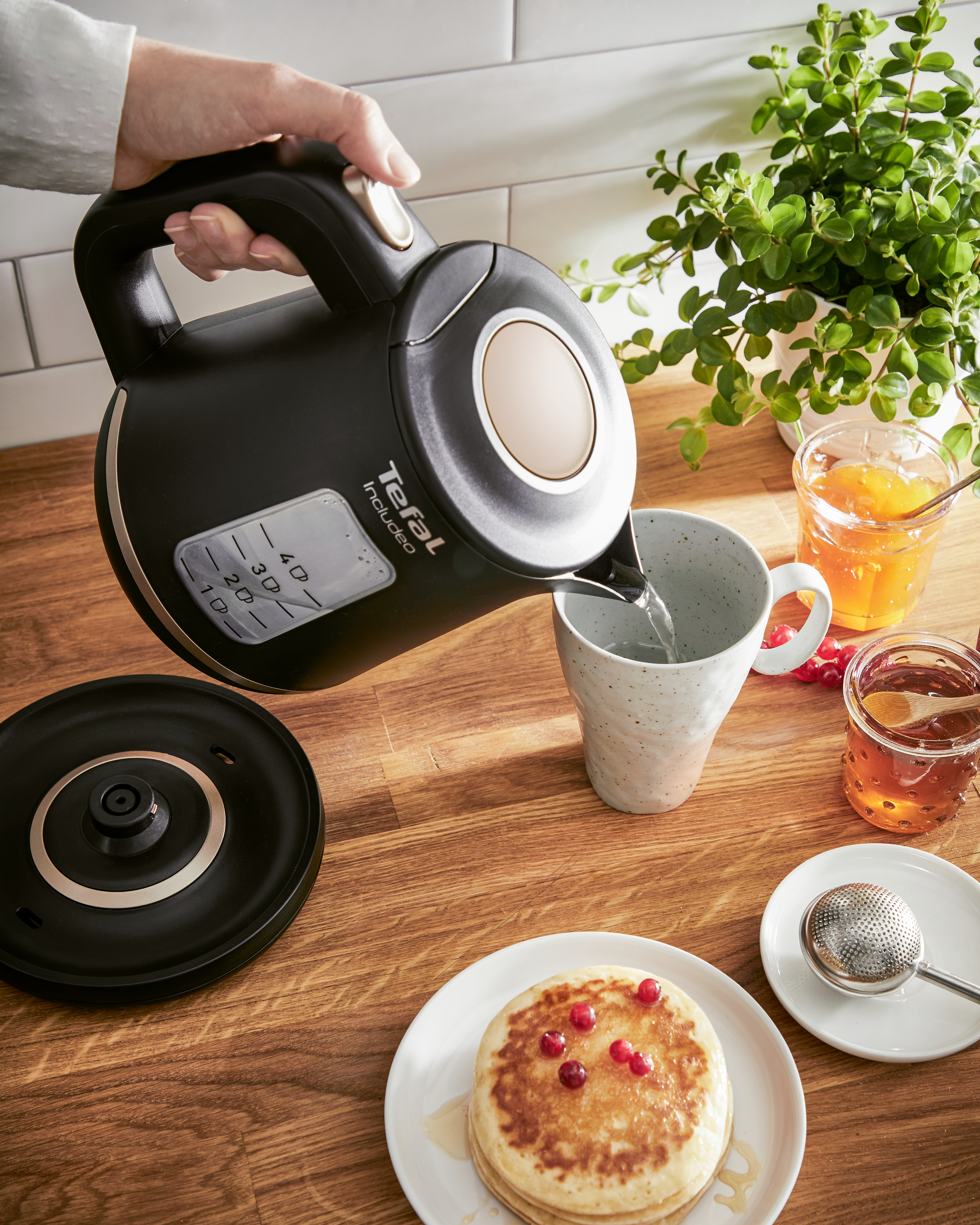 Eine Person gießt Wasser aus einem schwarzen Wasserkocher in eine Tasse mit Pfannkuchen und Tee.