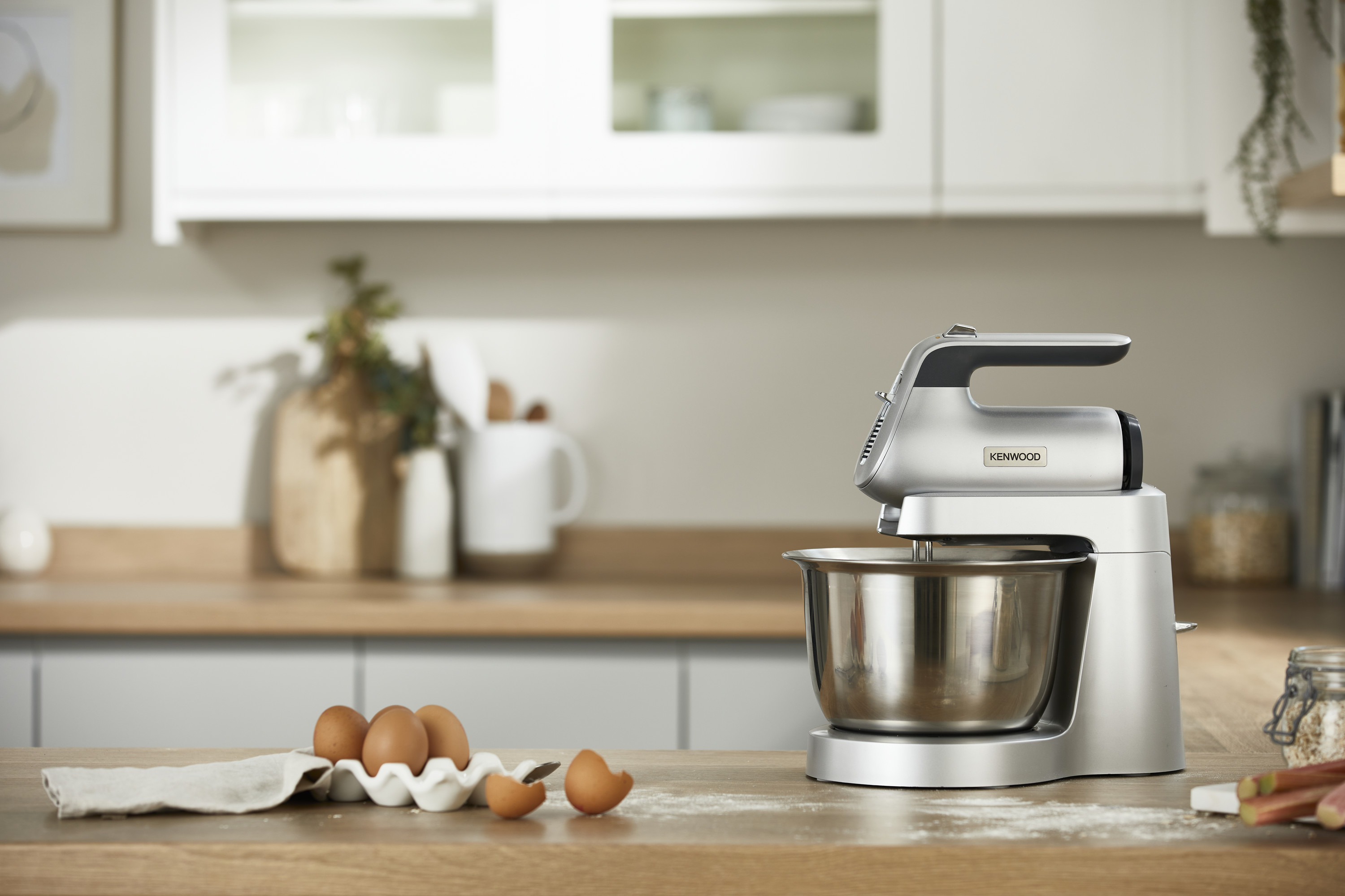 Ein silberner Kenwood Küchenmixer mit Schüssel, Eiern und Mehl auf einer Holztheke.