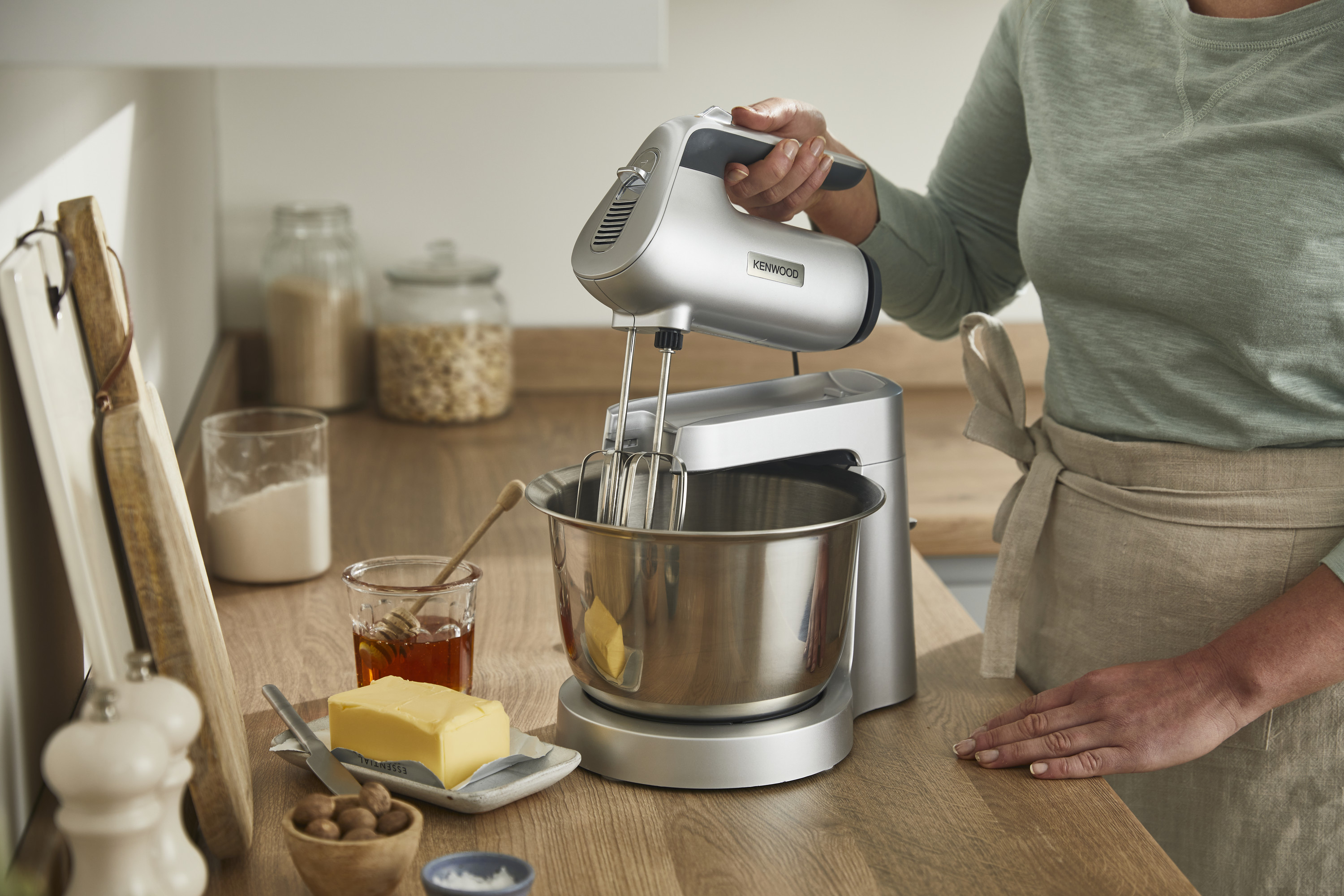 Eine Person benutzt einen silbernen Handmixer und eine Schüssel auf einer hölzernen Arbeitsplatte. Zutaten sichtbar.