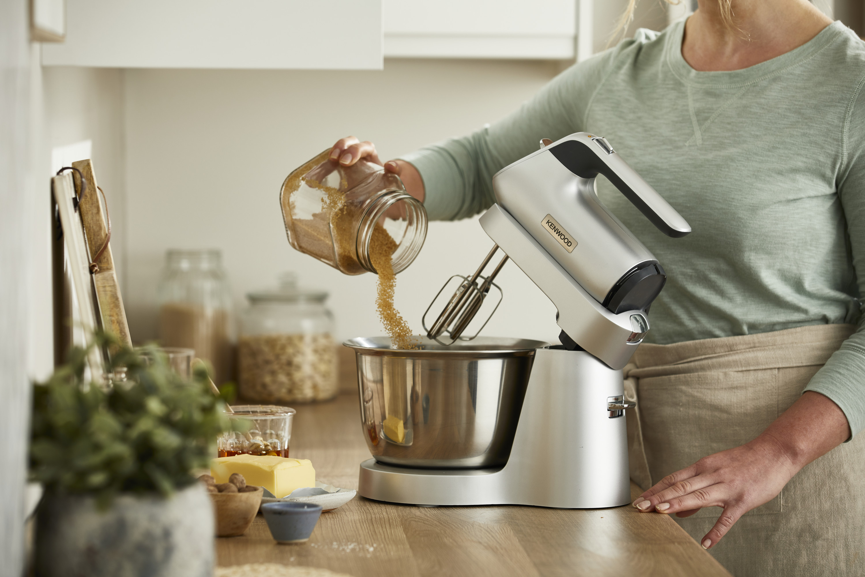 Eine Frau gießt braunen Zucker in eine Schüssel mit einem Standmixer auf einer Holz-Arbeitsplatte.