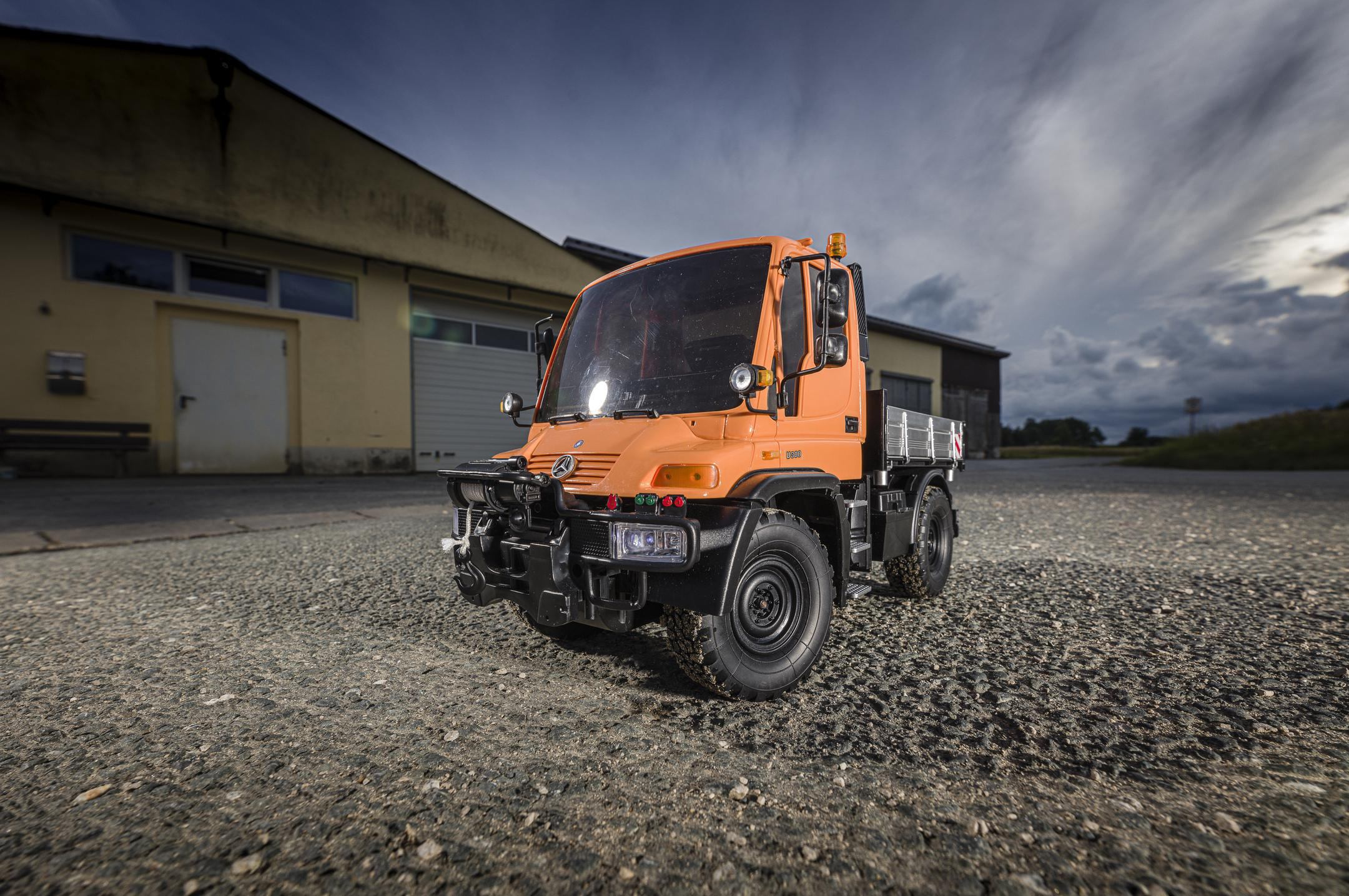 Oranger Unimog-Lkw auf einer Schotterstraße. Gebäude und ein bewölkter Himmel im Hintergrund.