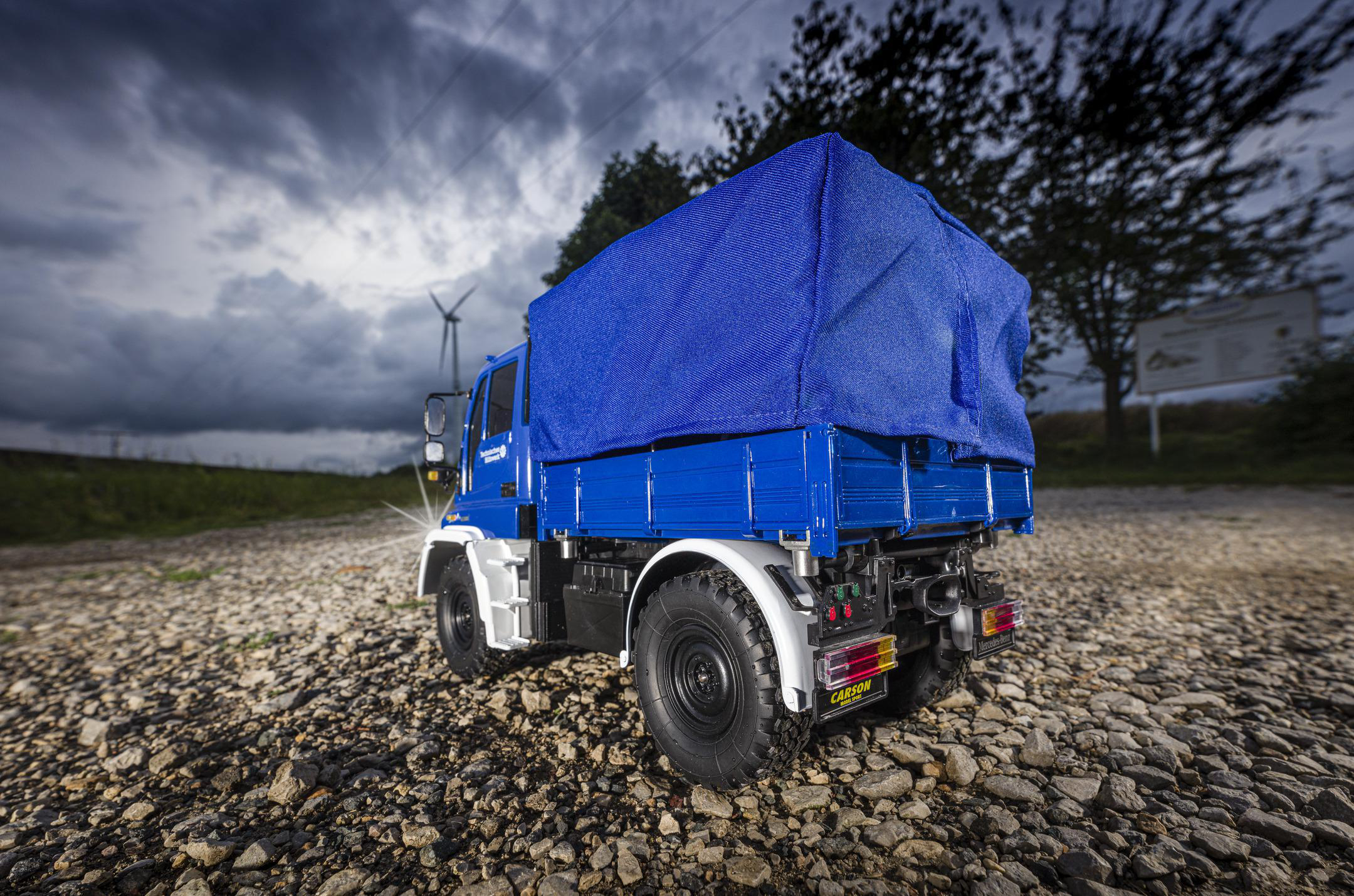 Ein blauer LKW mit blauem Planen in einem Feld. Der Hintergrund hat einen bewölkten Himmel und Windräder.