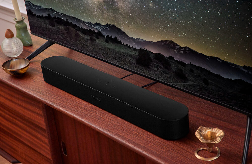 A black Sonos soundbar on a wooden table. A TV is behind it, with a mountain view.