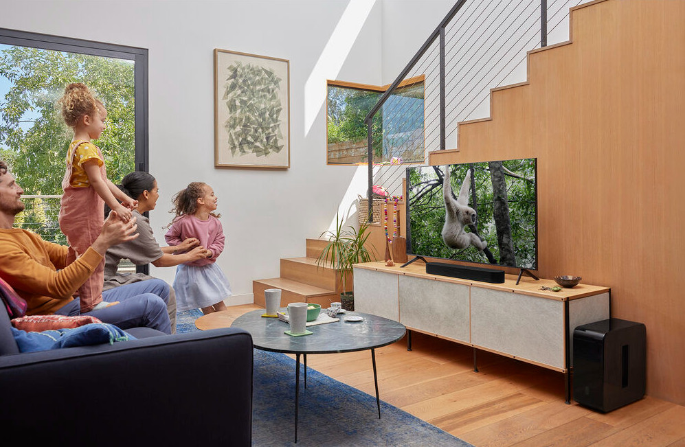 Family watches TV: parents, two kids, and a dog are in a living room with a TV and a sound bar.