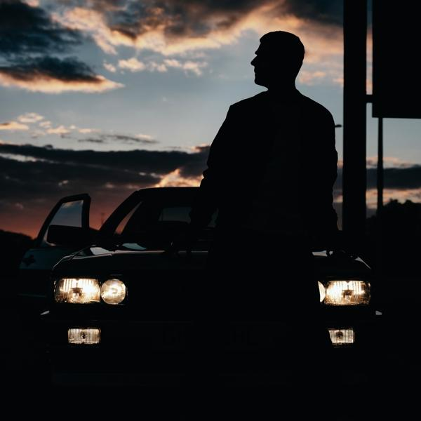 Silhouette d'un homme appuyé contre une voiture au crépuscule. Les phares sont allumés.