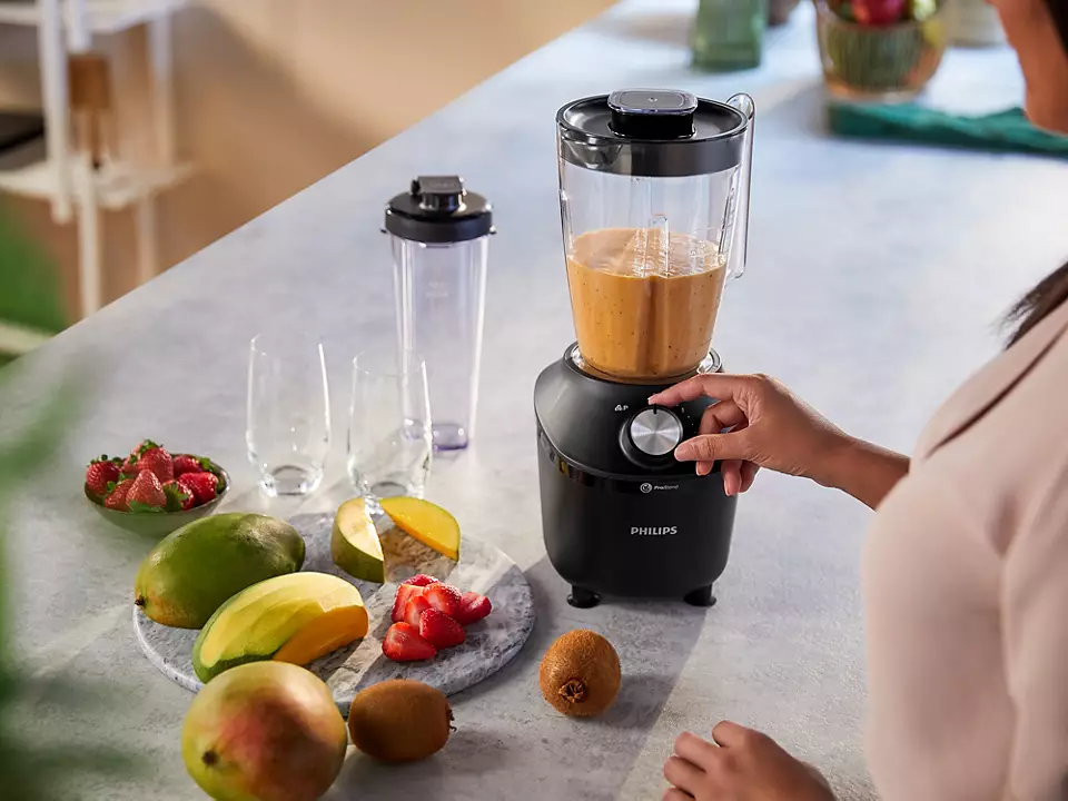 Eine Frau, die einen Mixer benutzt. Früchte und ein Smoothie sind sichtbar. Die Umgebung ist eine Küche.