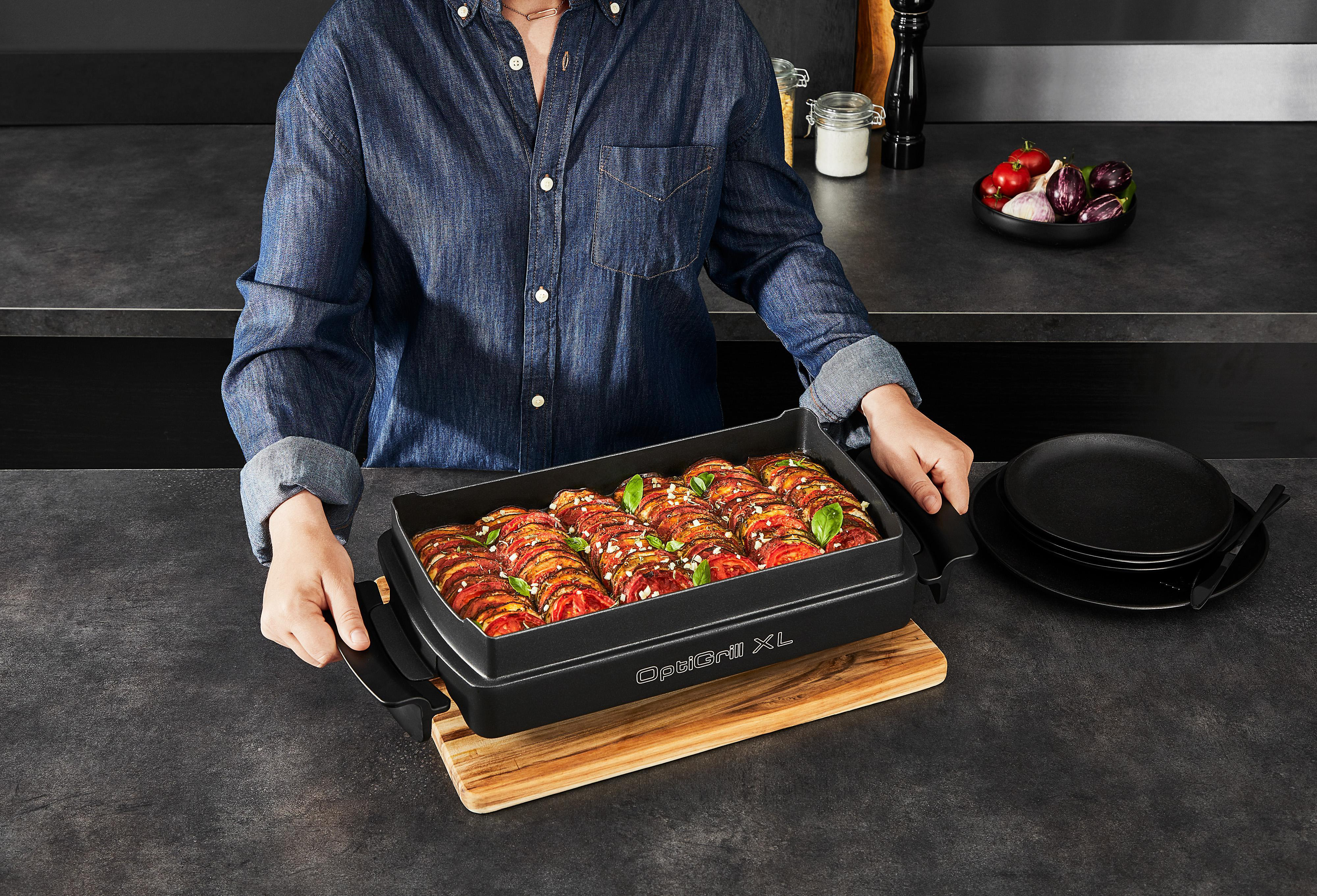 Person hält einen schwarzen OptiGrill XL mit gekochtem Essen auf einem Holzbrett.
