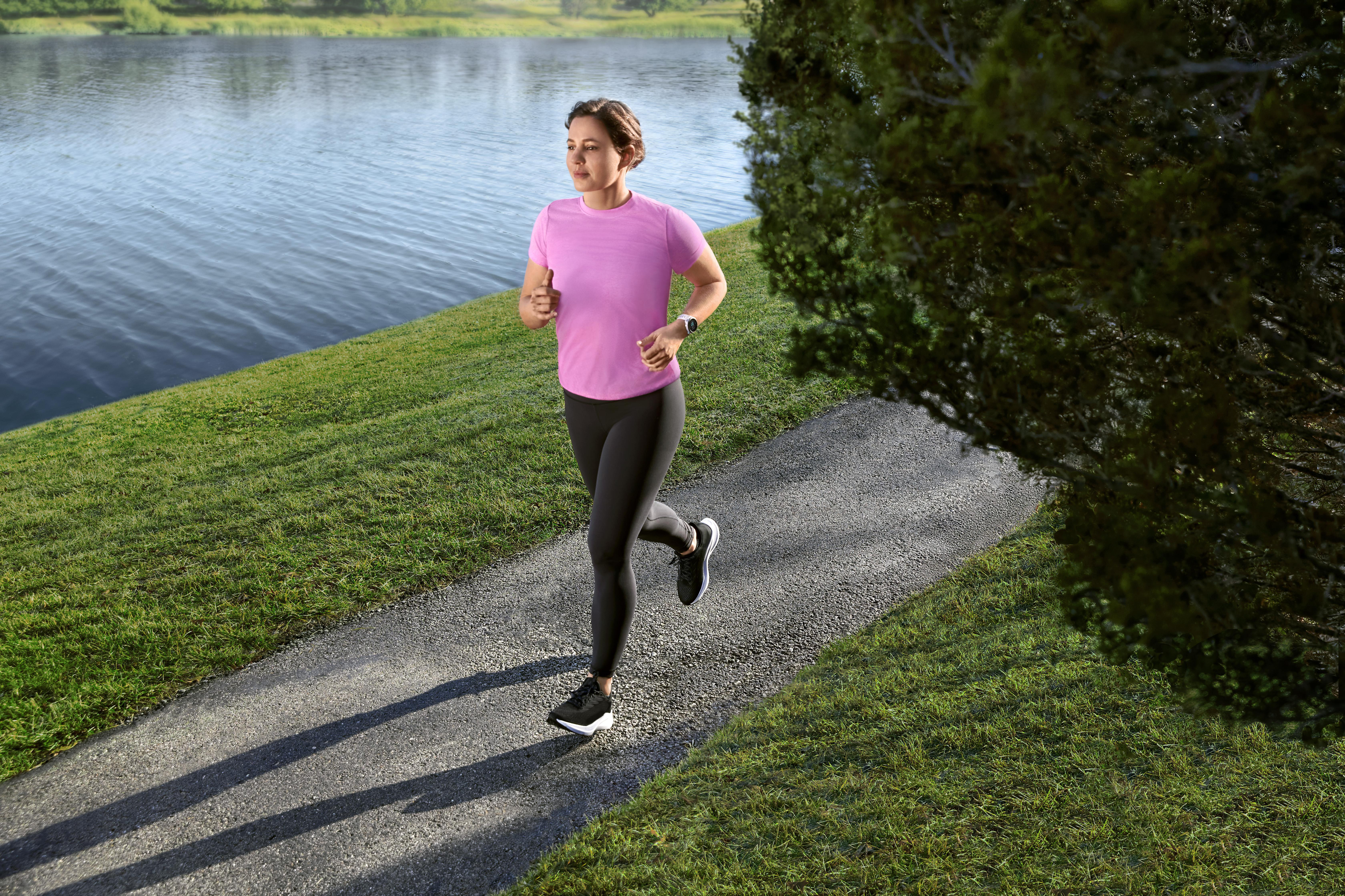 Eine Frau läuft auf einem Weg am See. Sie trägt ein pinkes Shirt und schwarze Leggings, mit grünem Gras daneben.