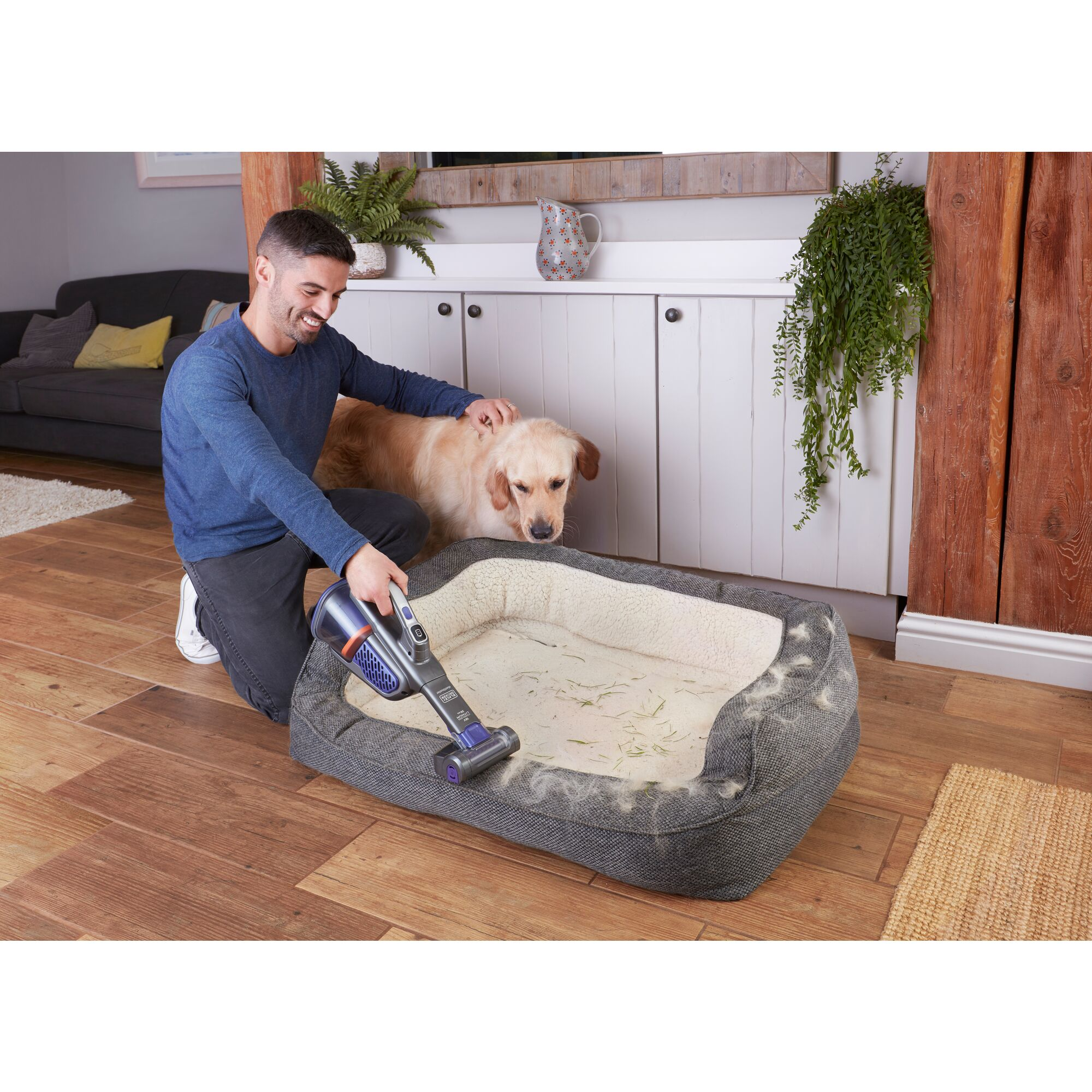 A man vacuums a dog bed while a dog is petted by him nearby.