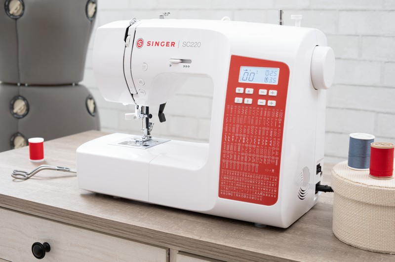 Weiße Nähmaschine auf einem Tisch, mit roten und blauen Garnspulen. Ein rotes Display ist sichtbar.