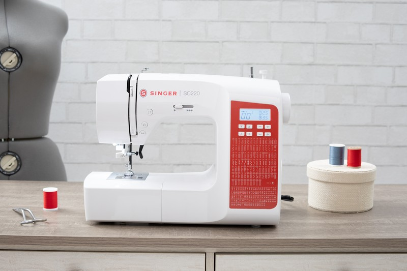 Eine weiße Singer SC220 Nähmaschine auf einem Holztisch mit Zubehör.