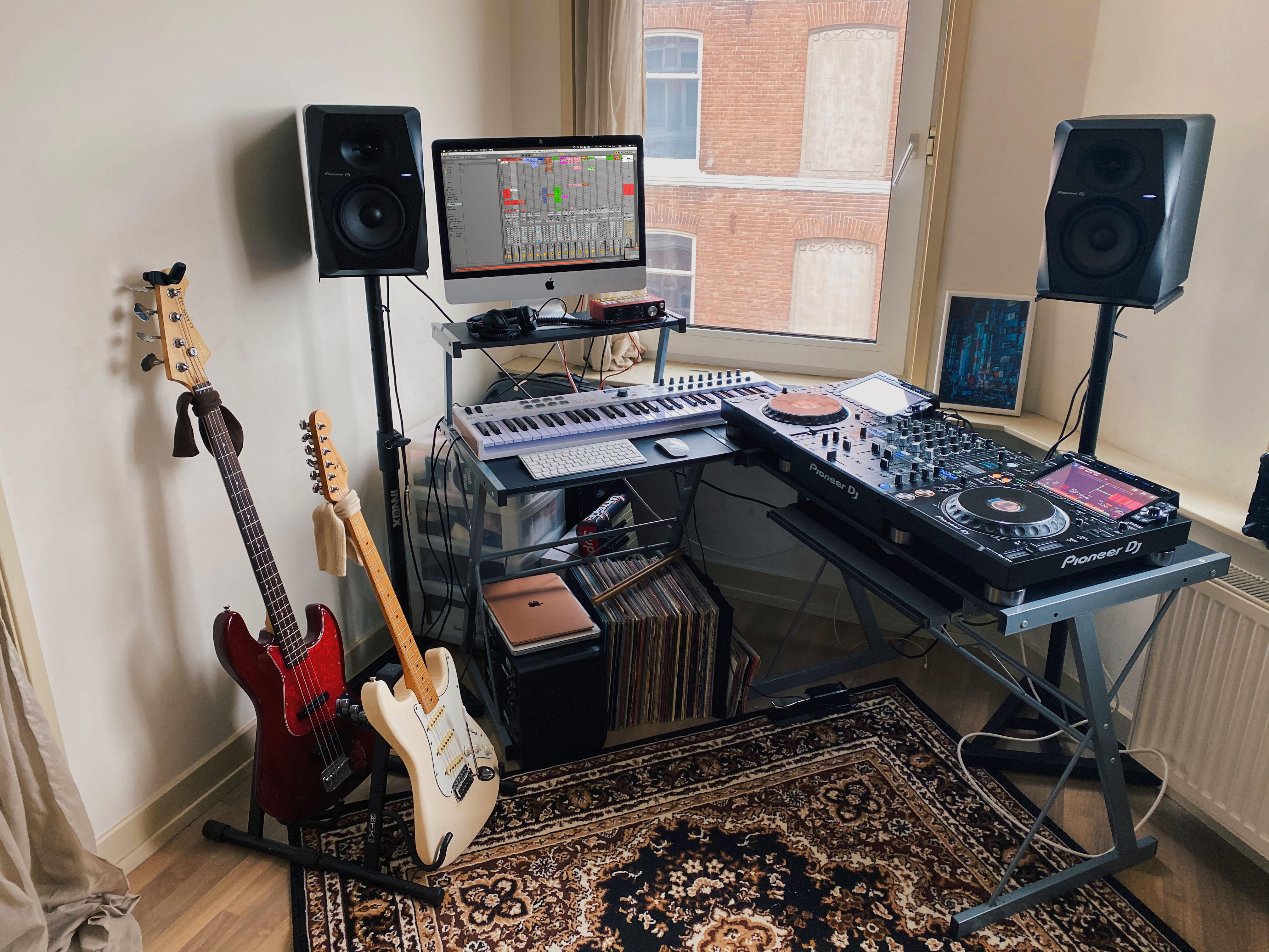 Ein Musikstudio mit Computer, Keyboard, DJ-Mixer, zwei Gitarren und Lautsprechern. Ein Fenster im Hintergrund.