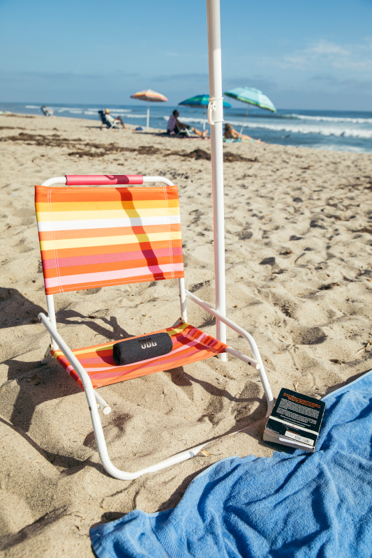 Scène de plage avec chaise rayée, haut-parleur, livre et parasol. Des gens sont en arrière-plan.