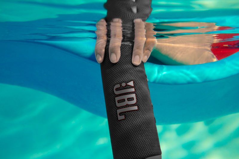 Main tenant un haut-parleur JBL noir dans l'eau, flotteur bleu en arrière-plan.