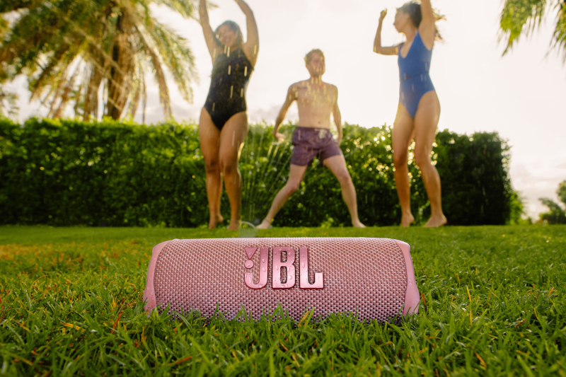 Enceinte JBL rose sur l'herbe. Personnes en maillots de bain, éclaboussant l'eau, journée ensoleillée.