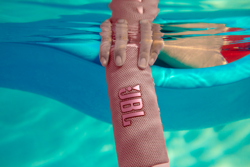 Haut-parleur JBL rose sous l'eau. Une main tient le haut-parleur. Dispositif de flottaison bleu en arrière-plan.