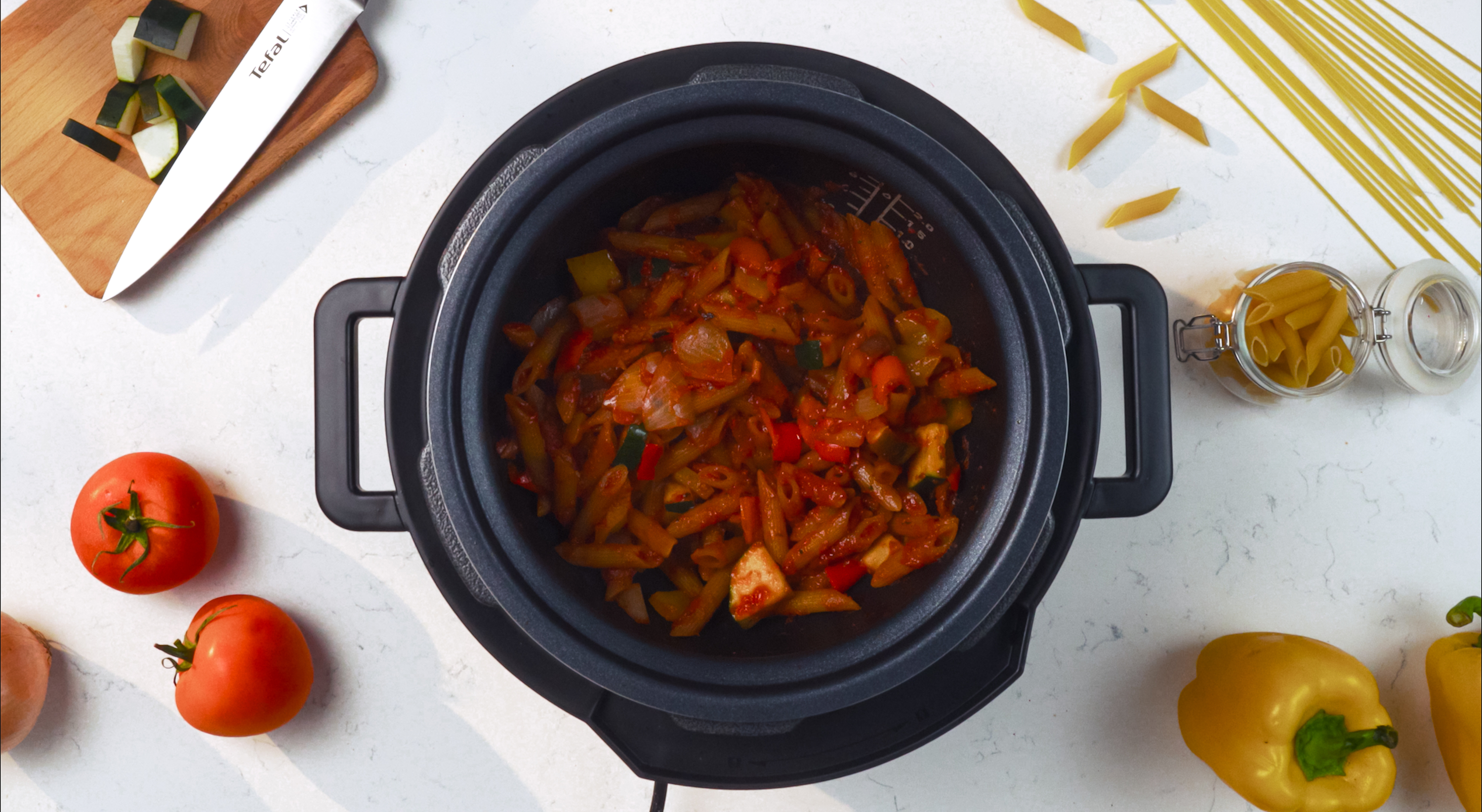 Pasta und Gemüse kochen in einem schwarzen Topf auf einer weißen Arbeitsplatte. Tomaten und Basilikum sind sichtbar.