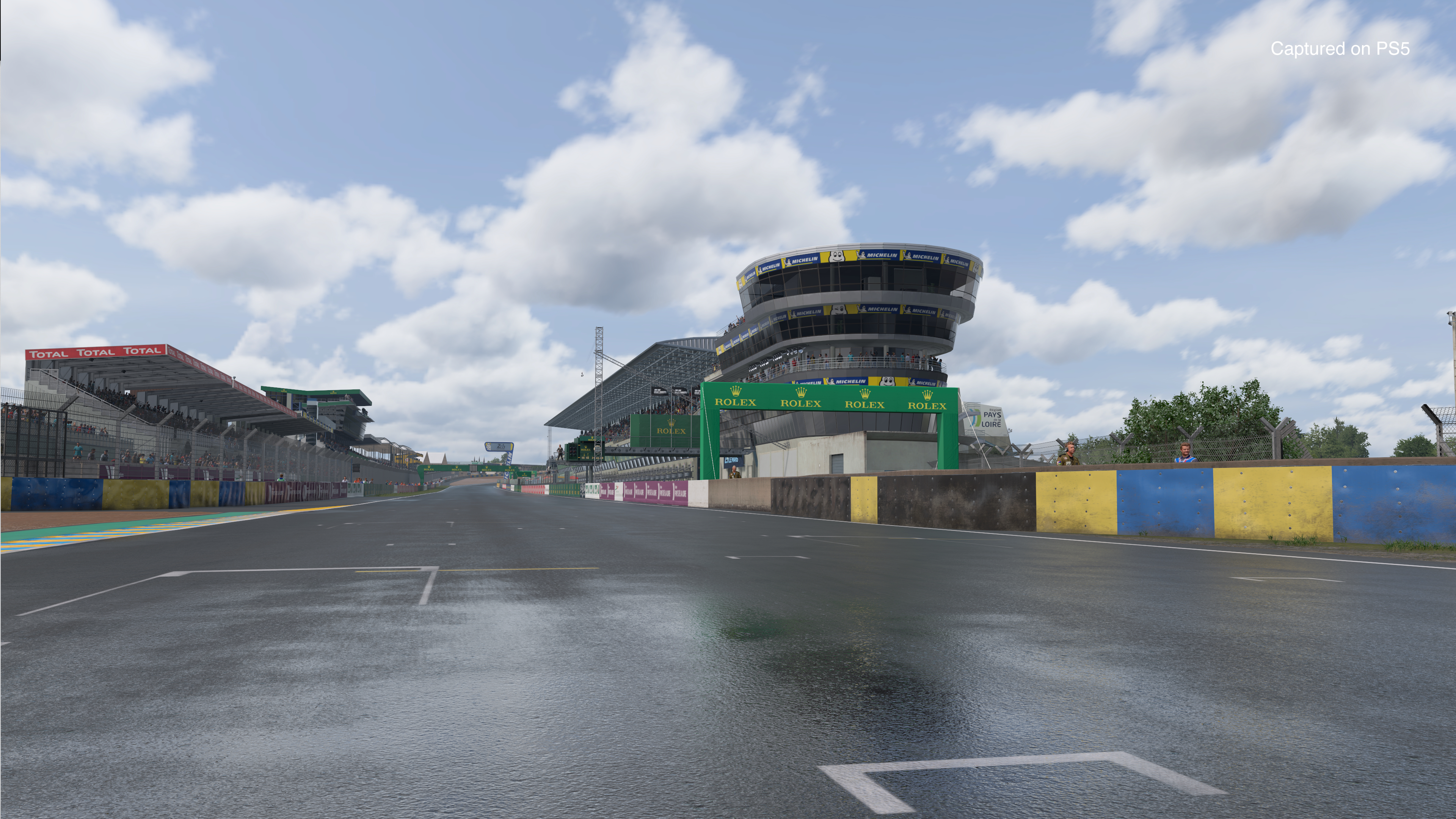 Rennstrecke in Le Mans mit Tribüne und Himmel. Beschilderung für Rolex und Total ist sichtbar.