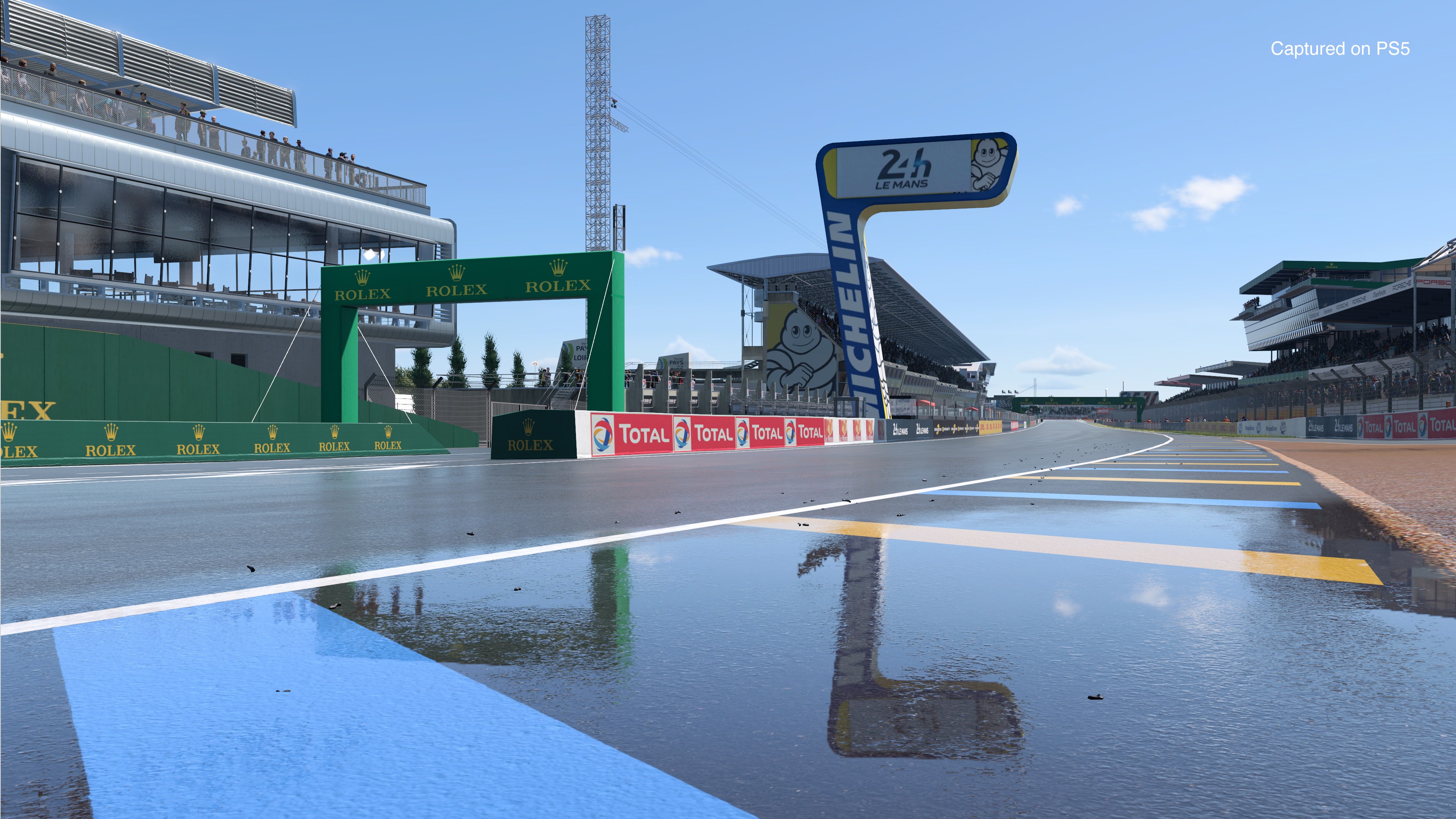 Une piste de course mouillée avec le panneau Le Mans, des bannières vertes et des tribunes sous un ciel bleu.