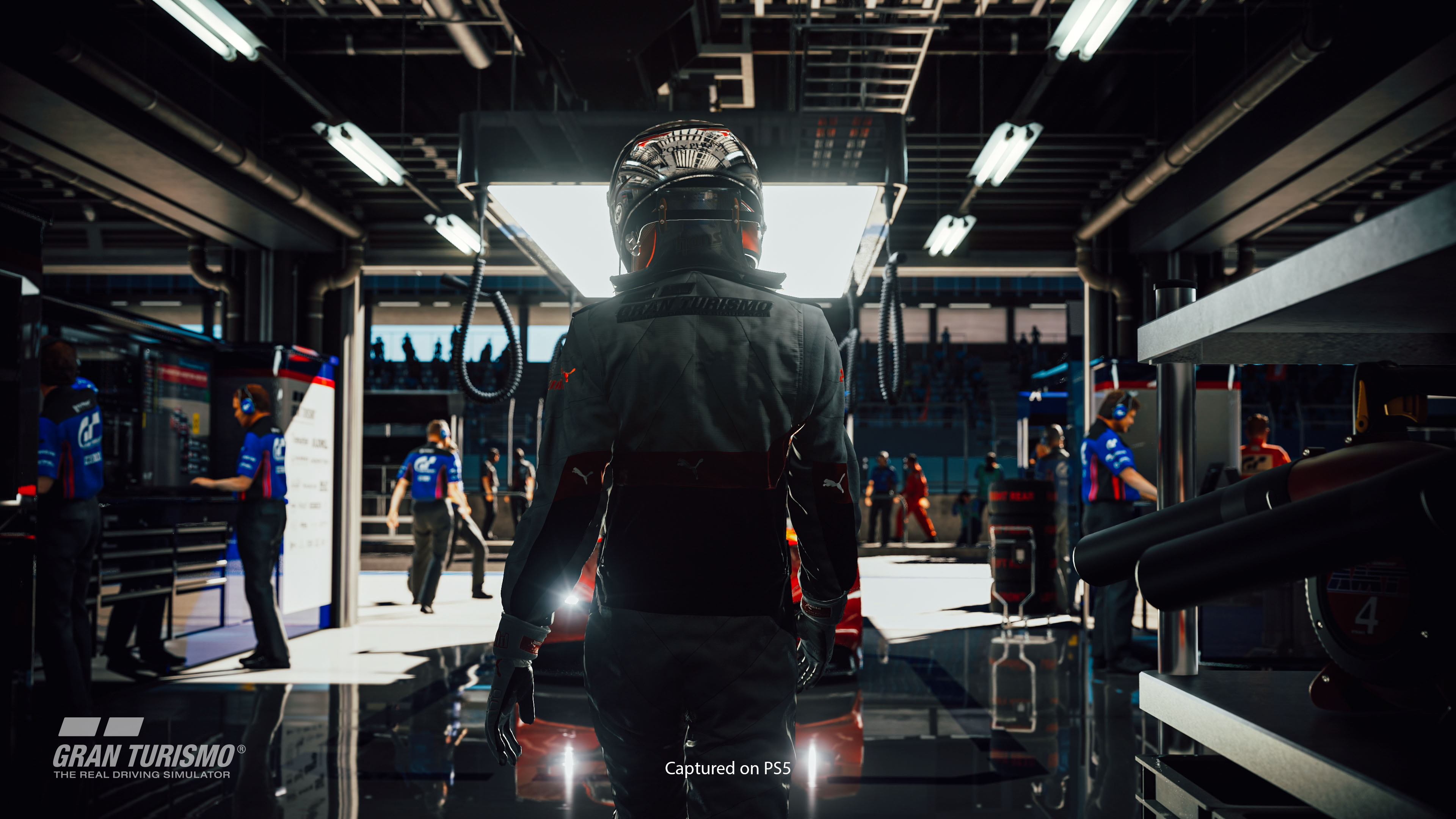 Une personne en tenue de course se tient dans un garage, regardant une voiture de course, logo Gran Turismo en dessous.