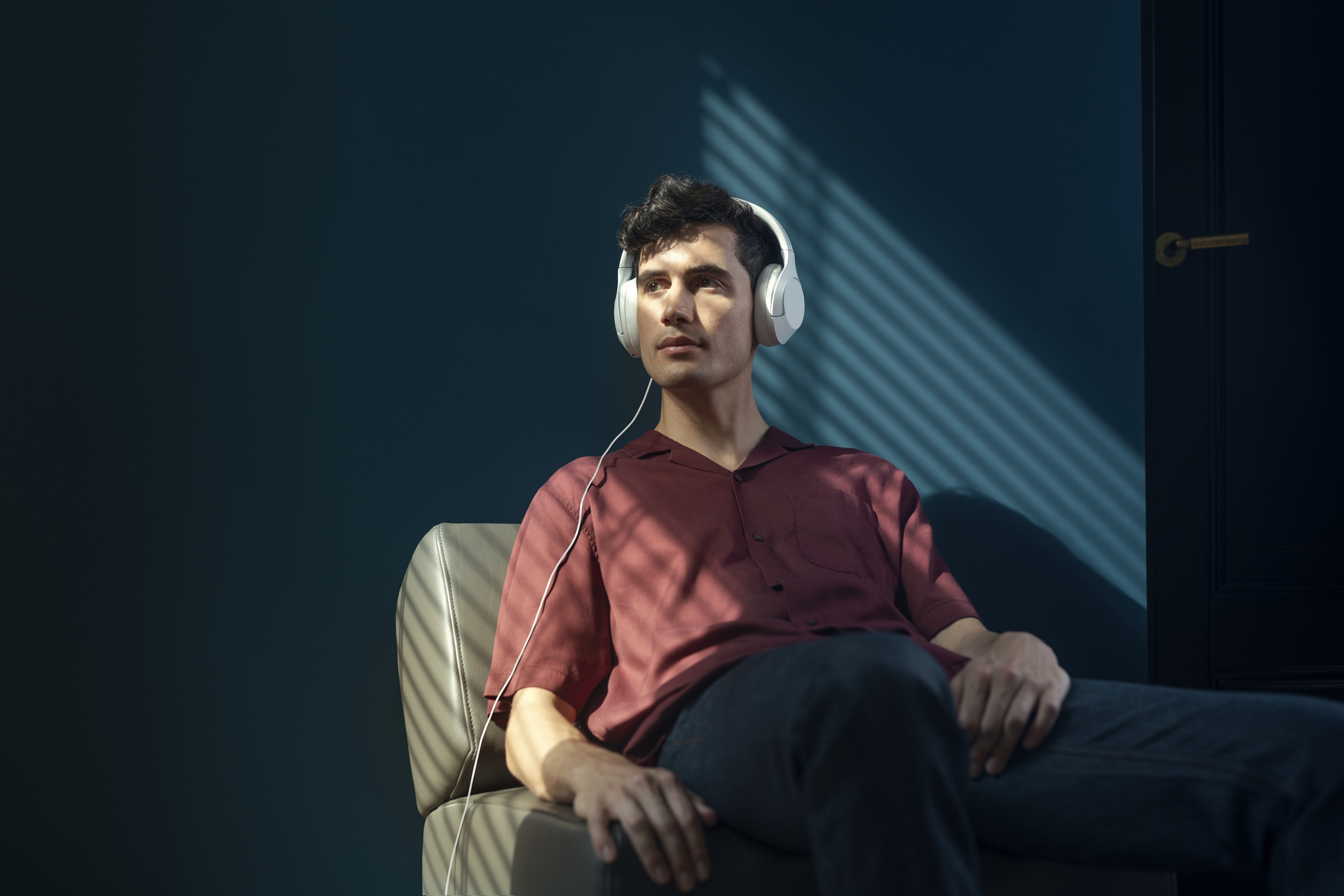 Mann mit weißen Kopfhörern, rotem Hemd und dunkler Hose, sitzt und blickt nach oben.