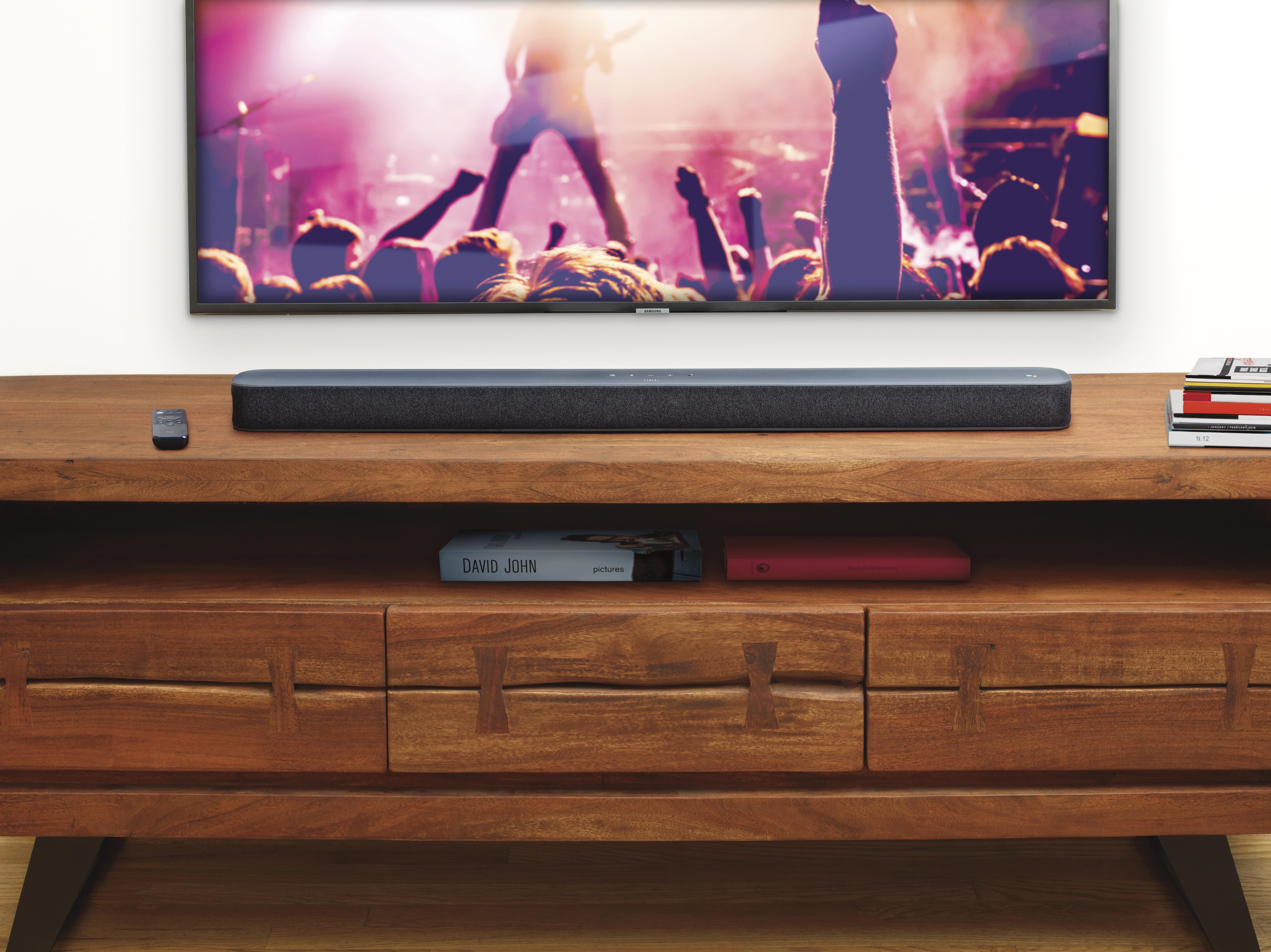 Fernseher auf einem Holzständer mit einer Soundbar darunter und einer Fernbedienung. Der Fernseher zeigt ein Konzert.