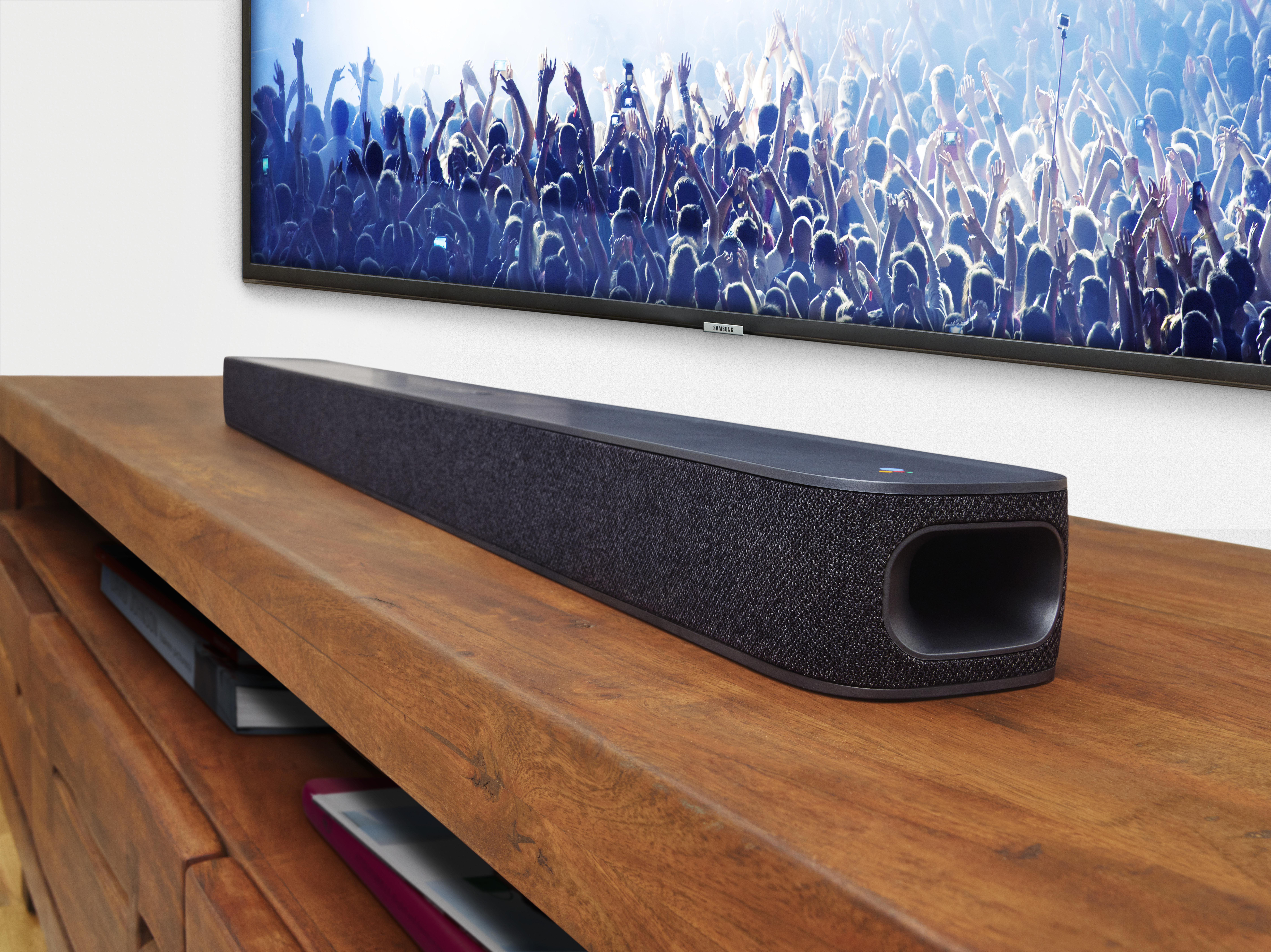 Soundbar auf einem Holzregal, mit einem Fernseher an der Wand, der eine Menschenmenge zeigt.