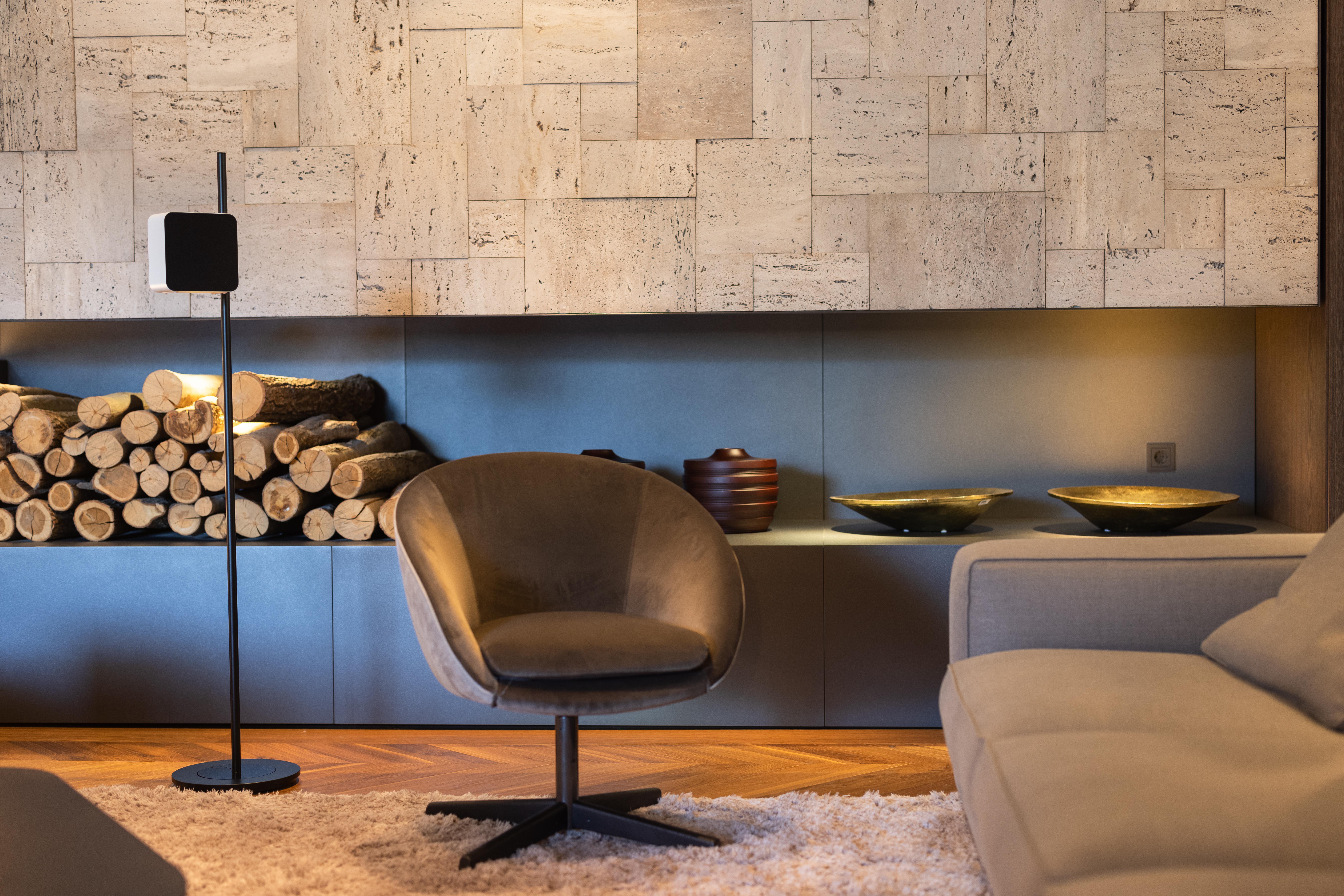 Ein modernes Wohnzimmer mit braunem Stuhl, Sofa und einem Stapel Holzscheite neben einer Steinmauer.