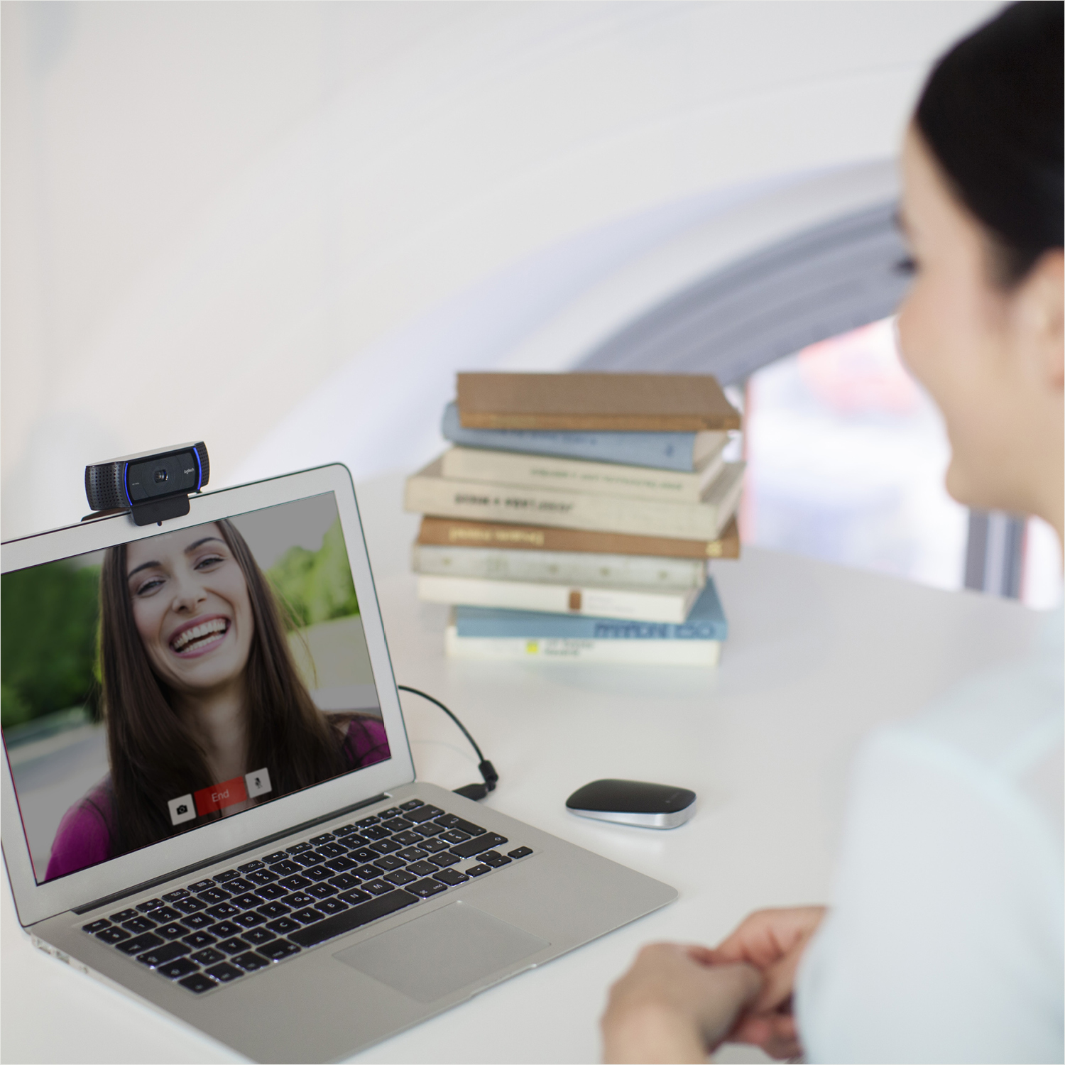 Frau bei einem Videoanruf, Laptop offen. Bücher und eine Maus liegen auf dem weißen Schreibtisch.