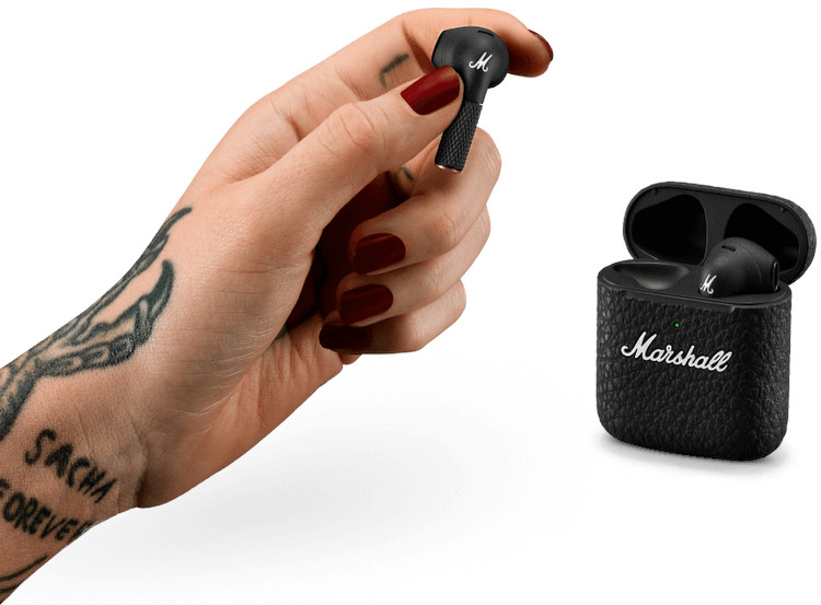 De hand van een persoon die een Marshall-oortelefoon en de oplaadcase vasthoudt. Getatoeëerde arm op de achtergrond.