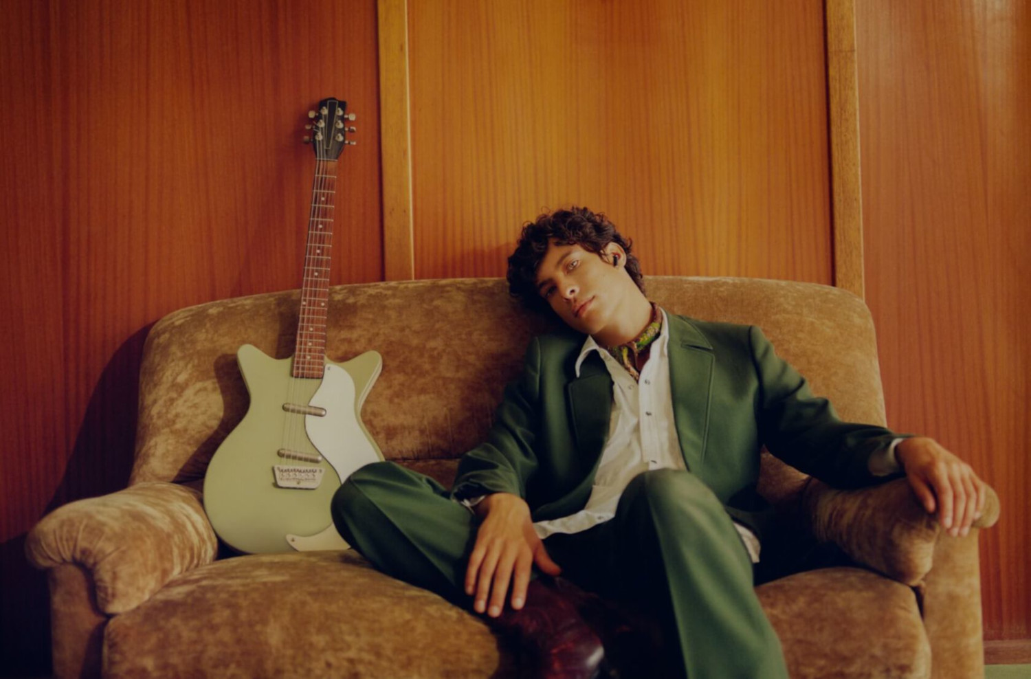Een man zit op een bruine bank met een gitaar. Hij draagt een groen pak.