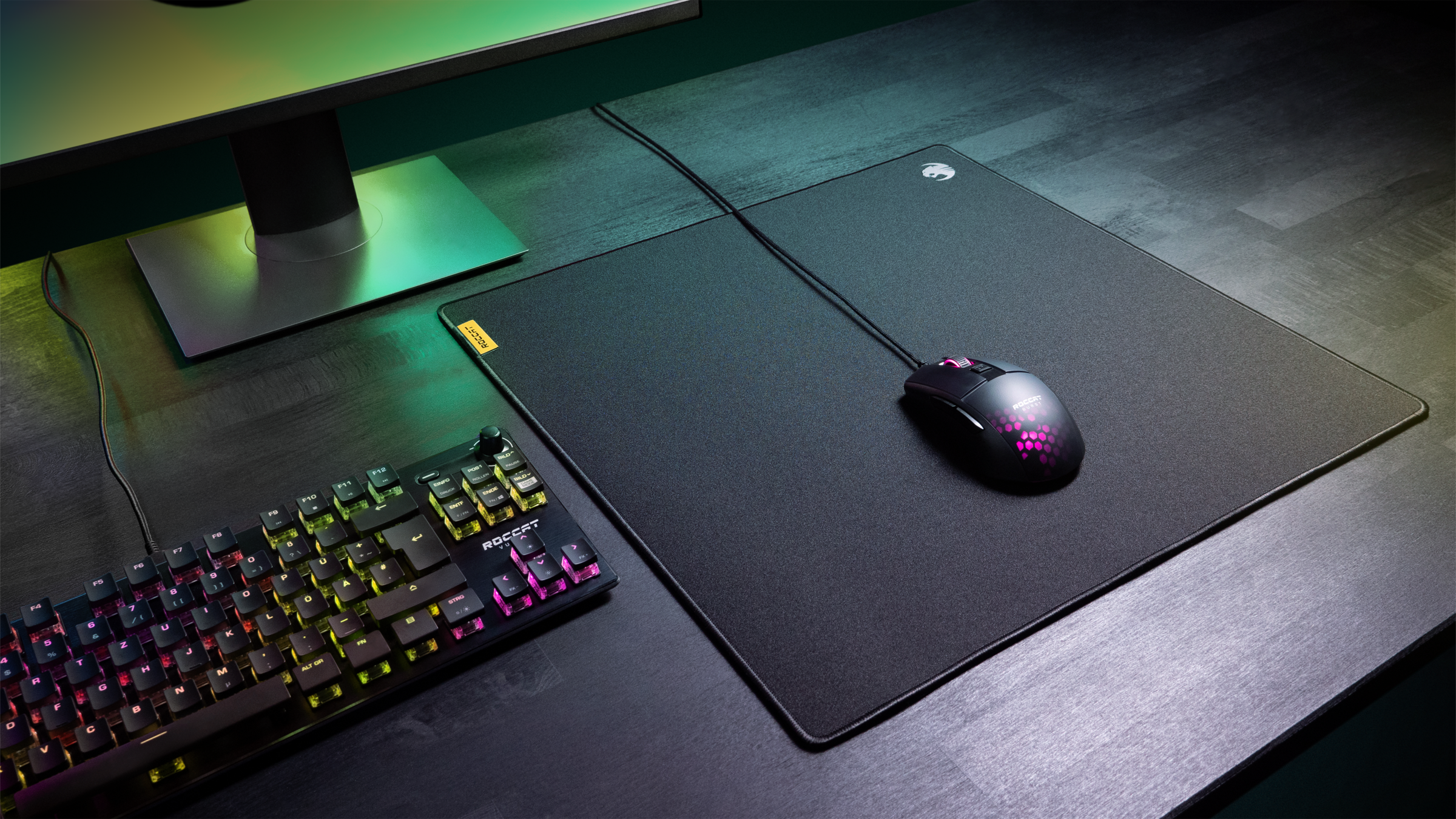 Gaming-Setup: Tastatur, Maus, Mauspad. Dunkler Schreibtisch, Monitor im Hintergrund.