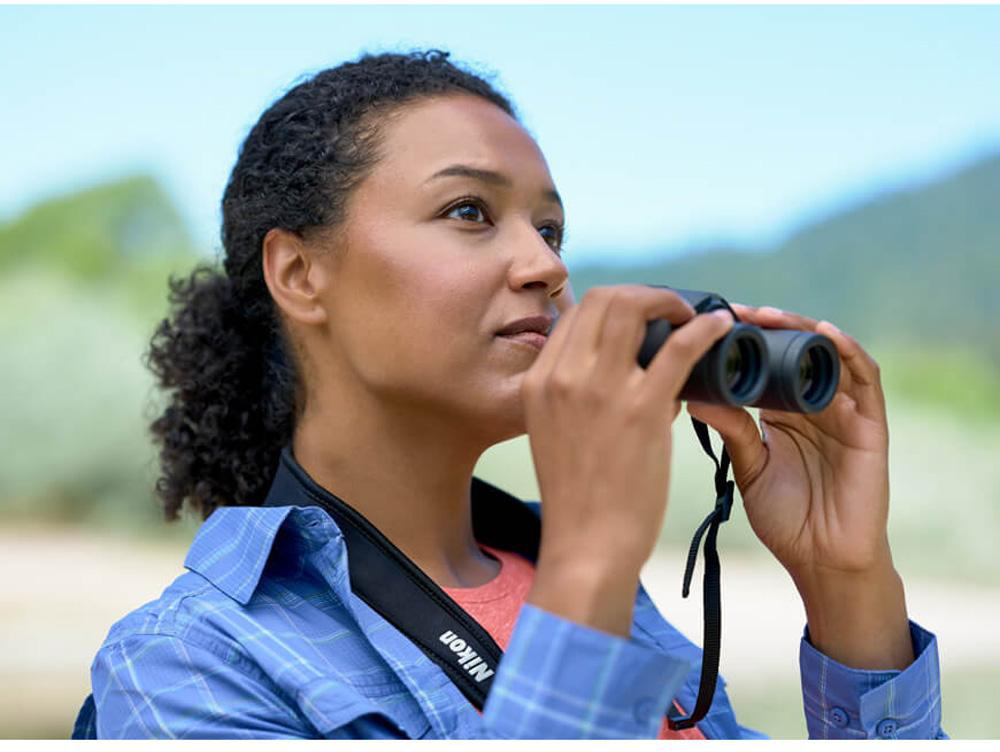 Donna che guarda attraverso un binocolo. È all'aperto, con alberi e spiaggia sullo sfondo.