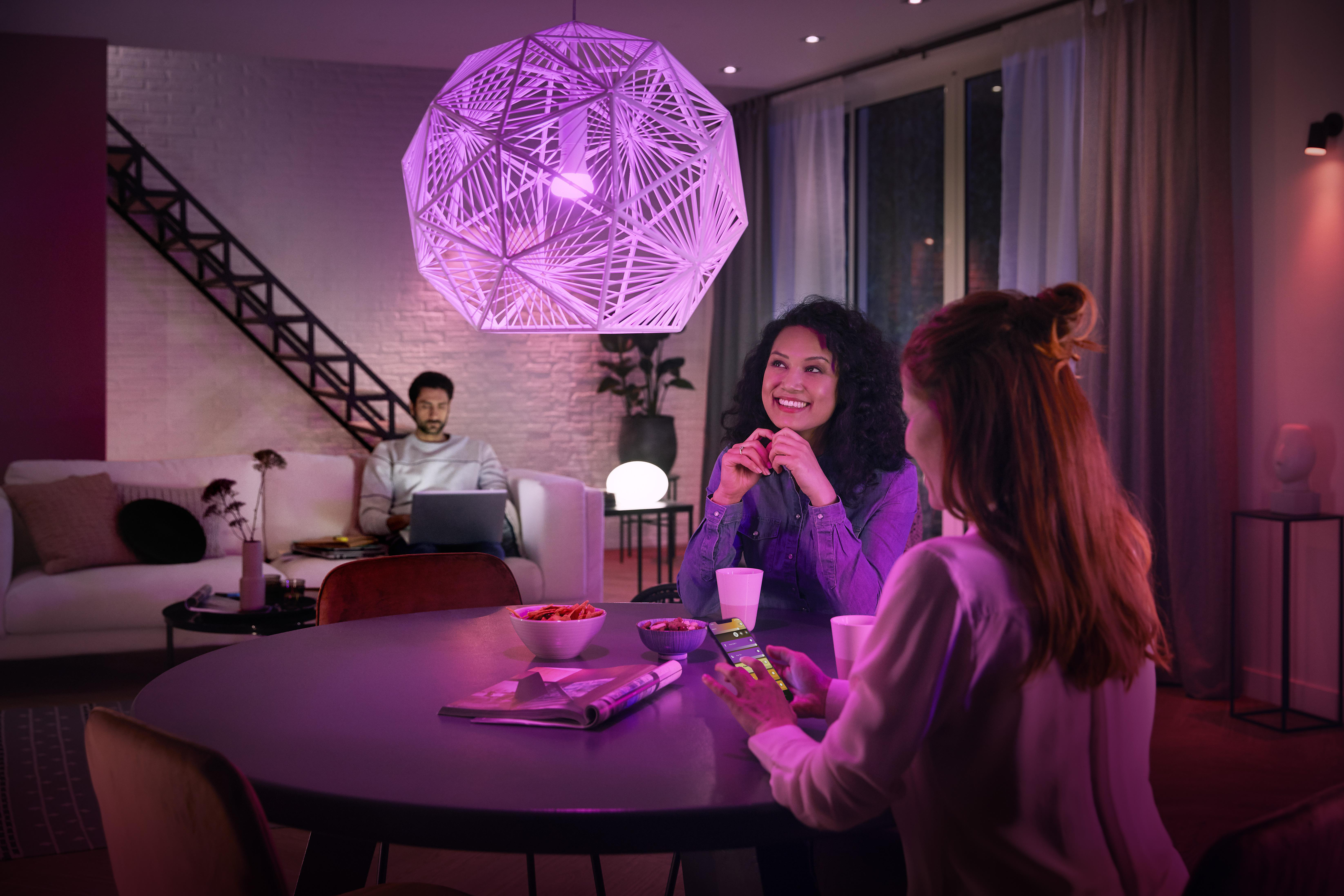 Zwei Frauen und ein Mann sitzen an einem Tisch. Lila Licht beleuchtet die Szene.
