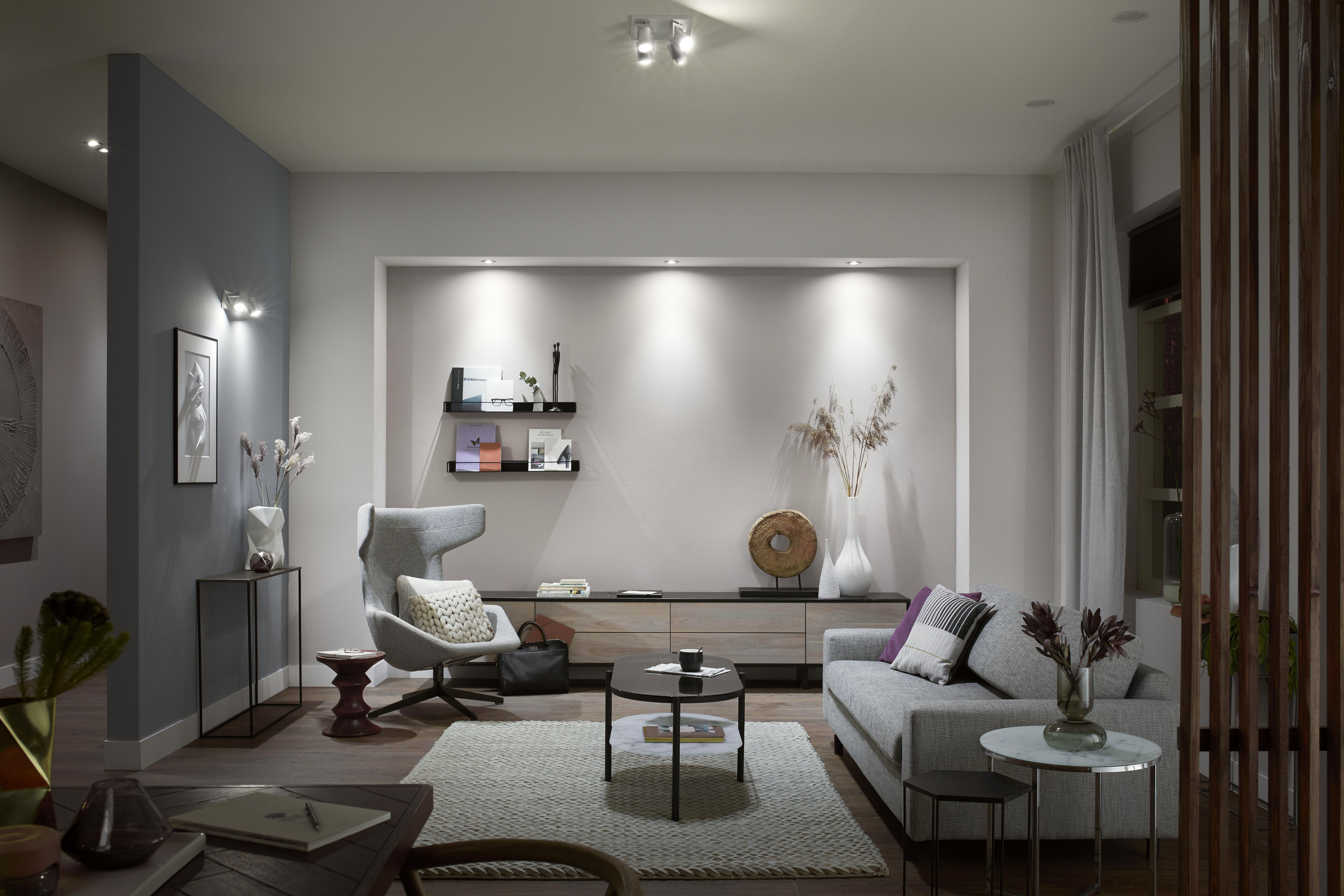 Wohnzimmer mit grauem Sofa, Holzschrank, Sessel und Couchtisch, gut beleuchtet durch Lampen.