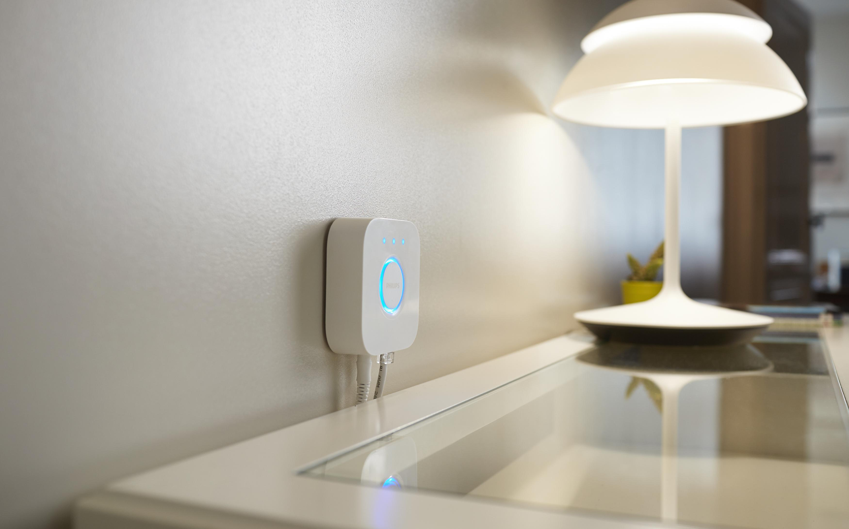 Ein weißer Smart Hub an einer weißen Wand. Daneben eine Tischlampe. Kabel sind sichtbar.