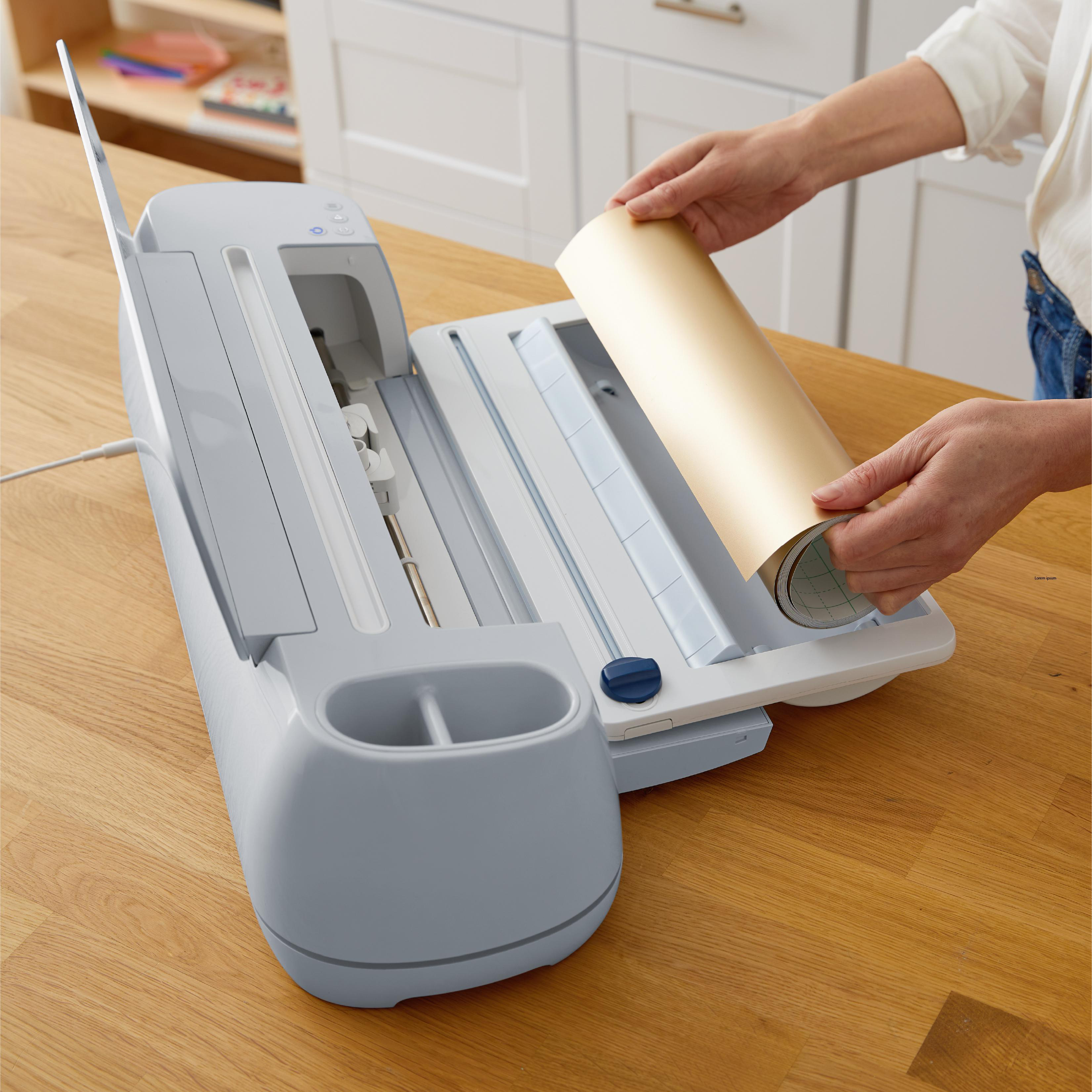 Eine Person legt eine goldene Materialrolle in eine Cricut-Maschine ein. Die Maschine steht auf einem Holztisch.