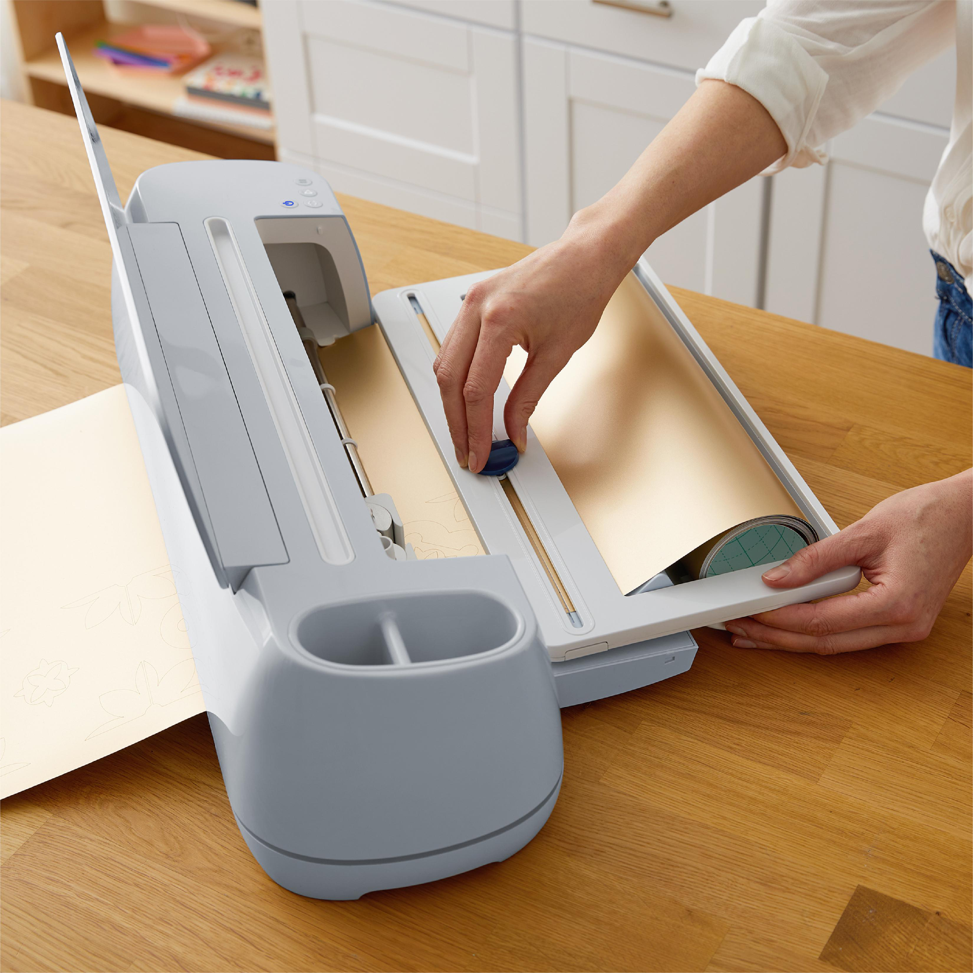Eine Person bedient eine Cricut-Maschine. Ein goldenes Blatt wird eingesetzt. Die Maschine steht auf einem Holztisch.