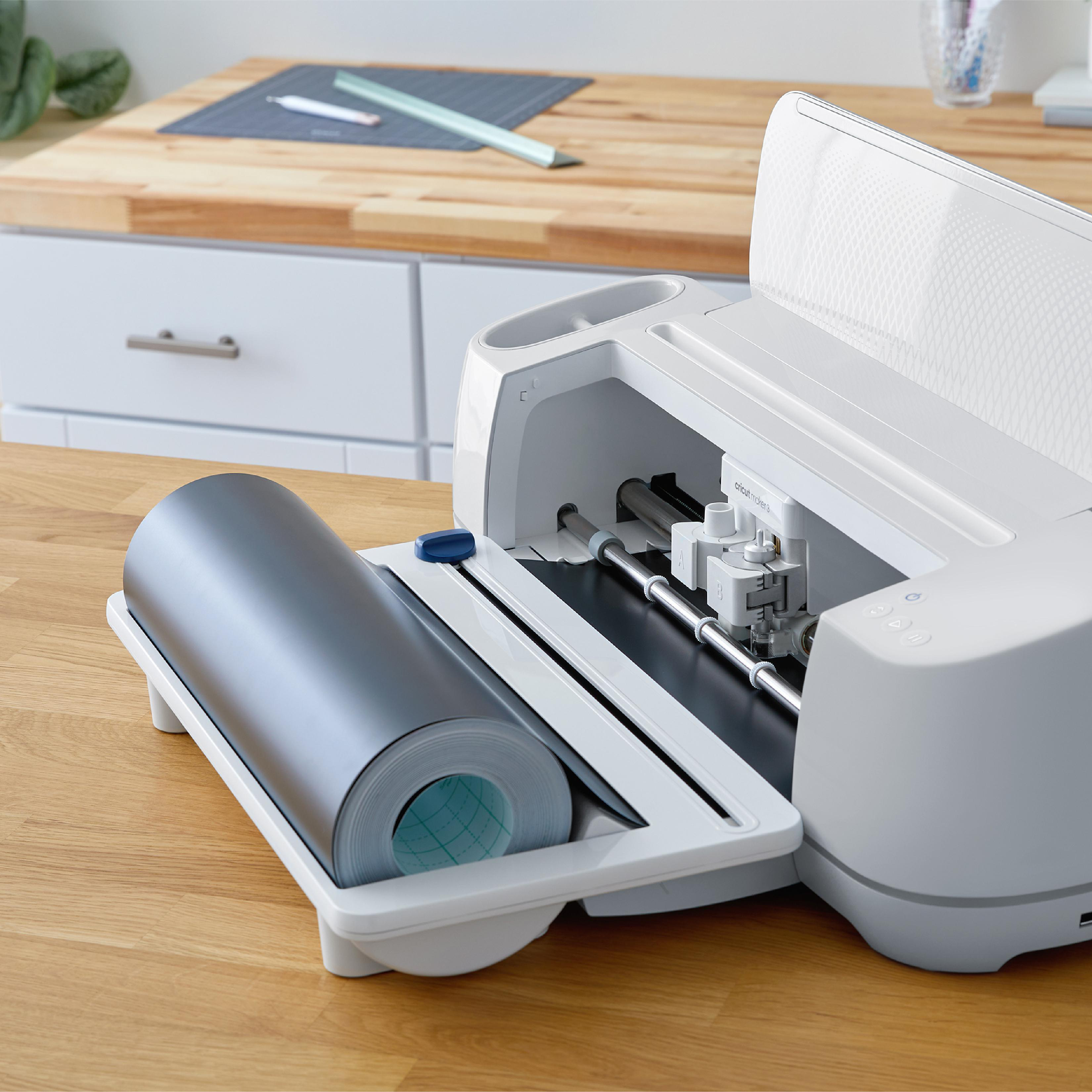 Eine weiße Cricut Maker 3 Maschine mit einer Rolle grauen Vinyls. Sie steht auf einer Holzoberfläche mit Bastelmaterialien.