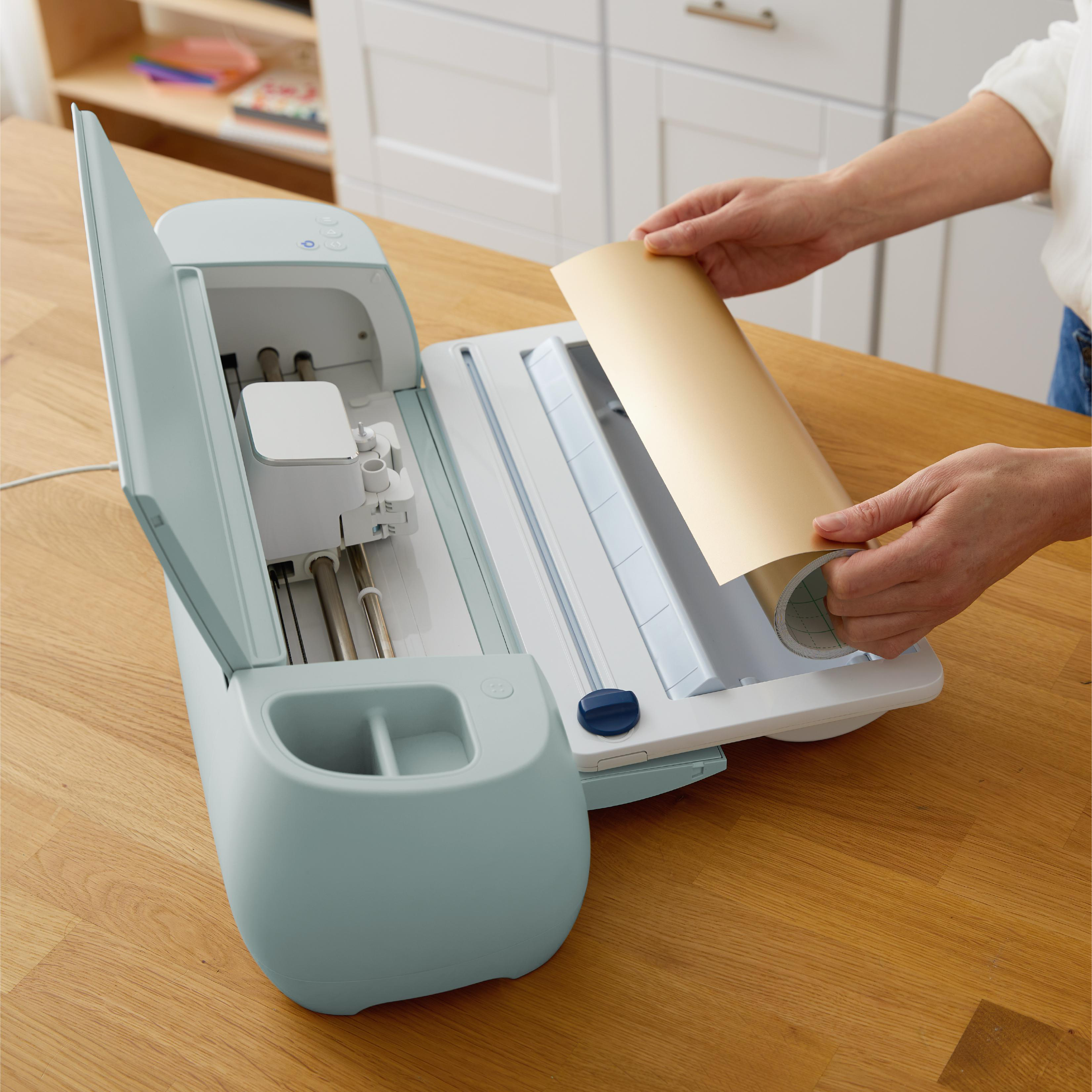 Eine Person bedient eine Schneidemaschine mit einer goldenen Rolle. Die Maschine ist hellblau. Sie steht auf einem Holztisch.