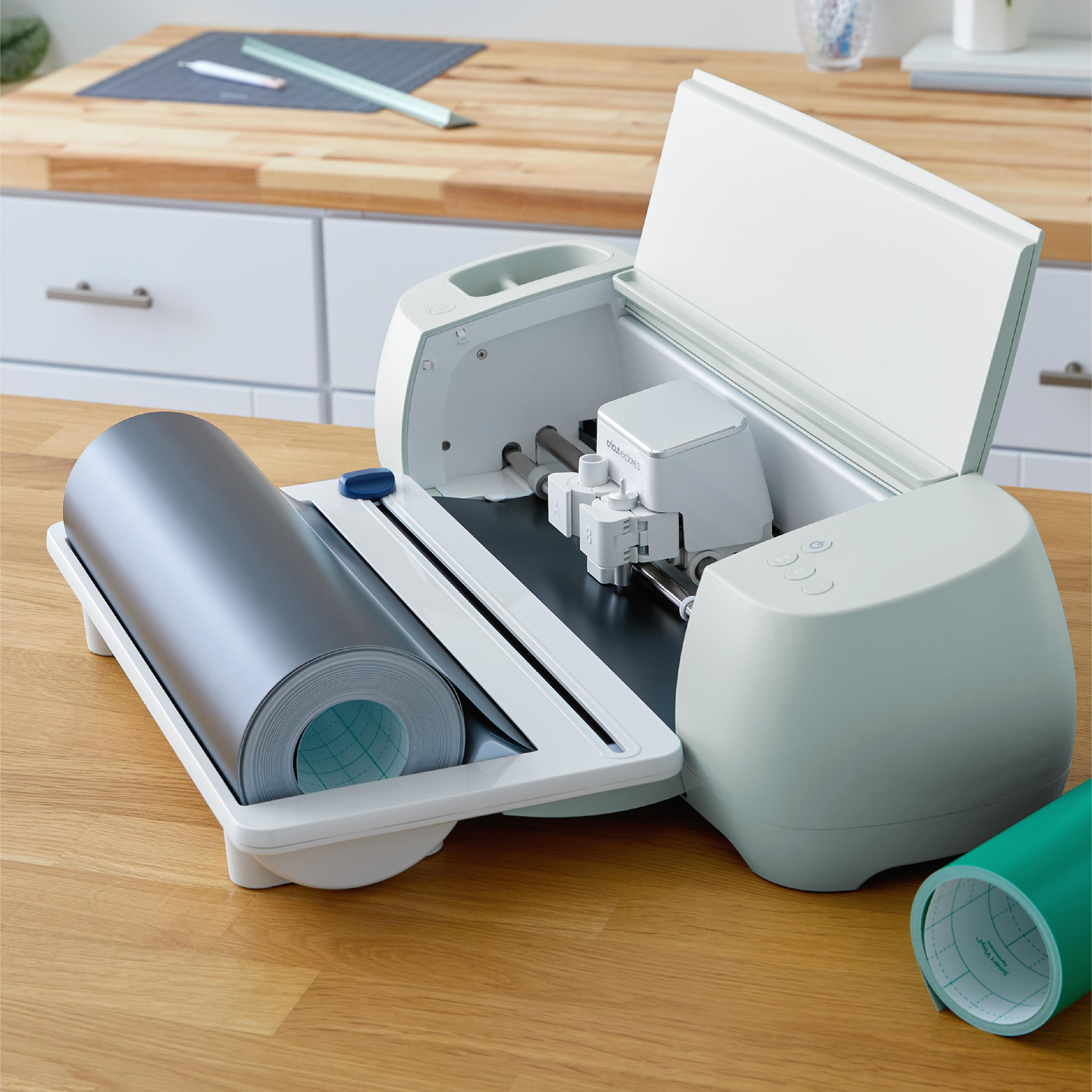 Eine Cricut-Maschine mit einer Rolle grauen Vinyls. Auf dem Holztisch befindet sich eine weitere grüne Rolle. Weiße Schränke im Hintergrund.