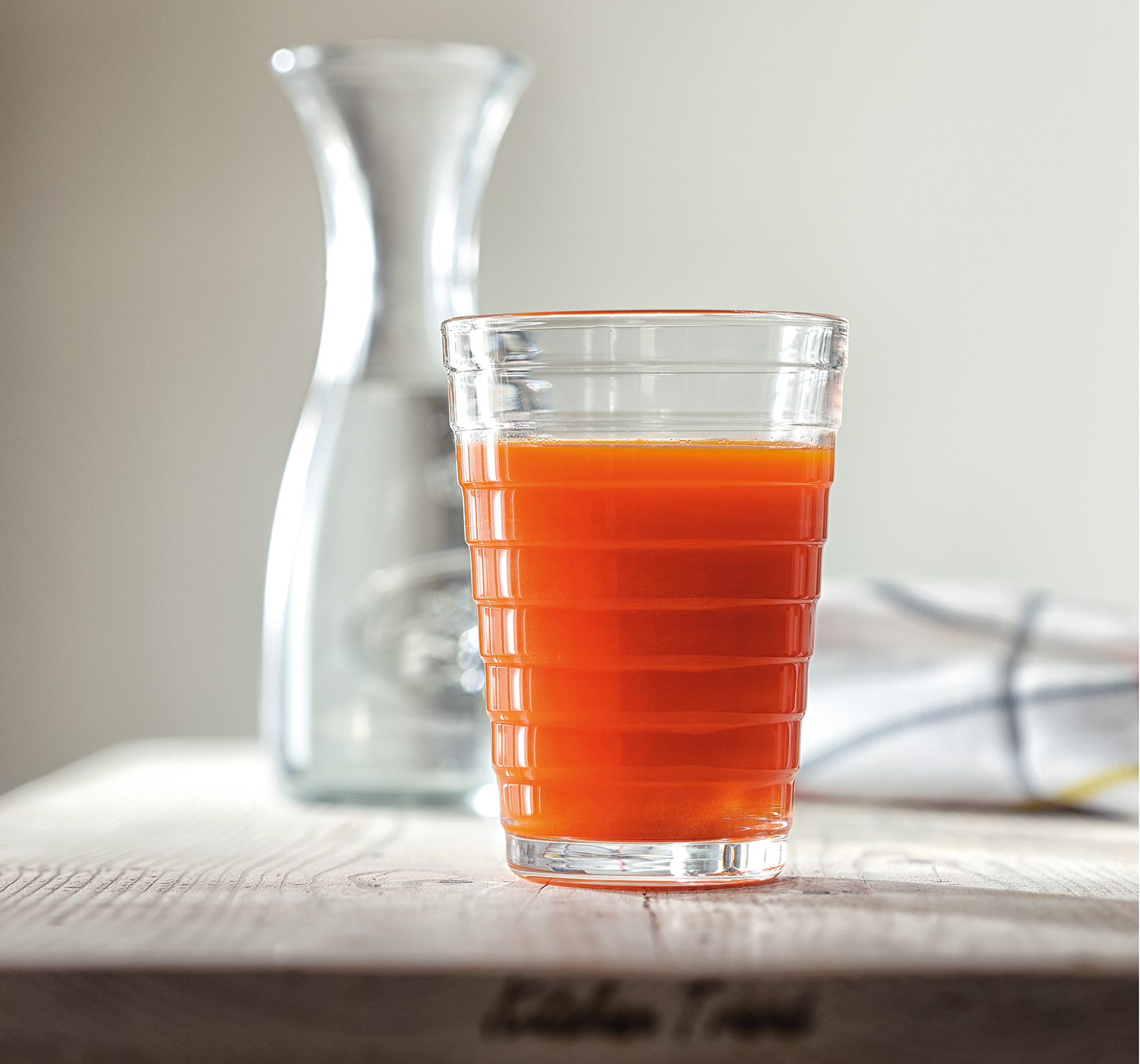 Een glas sinaasappelsap staat op een houten tafel. Een doorzichtige karaf en een doek met patroon op de achtergrond.