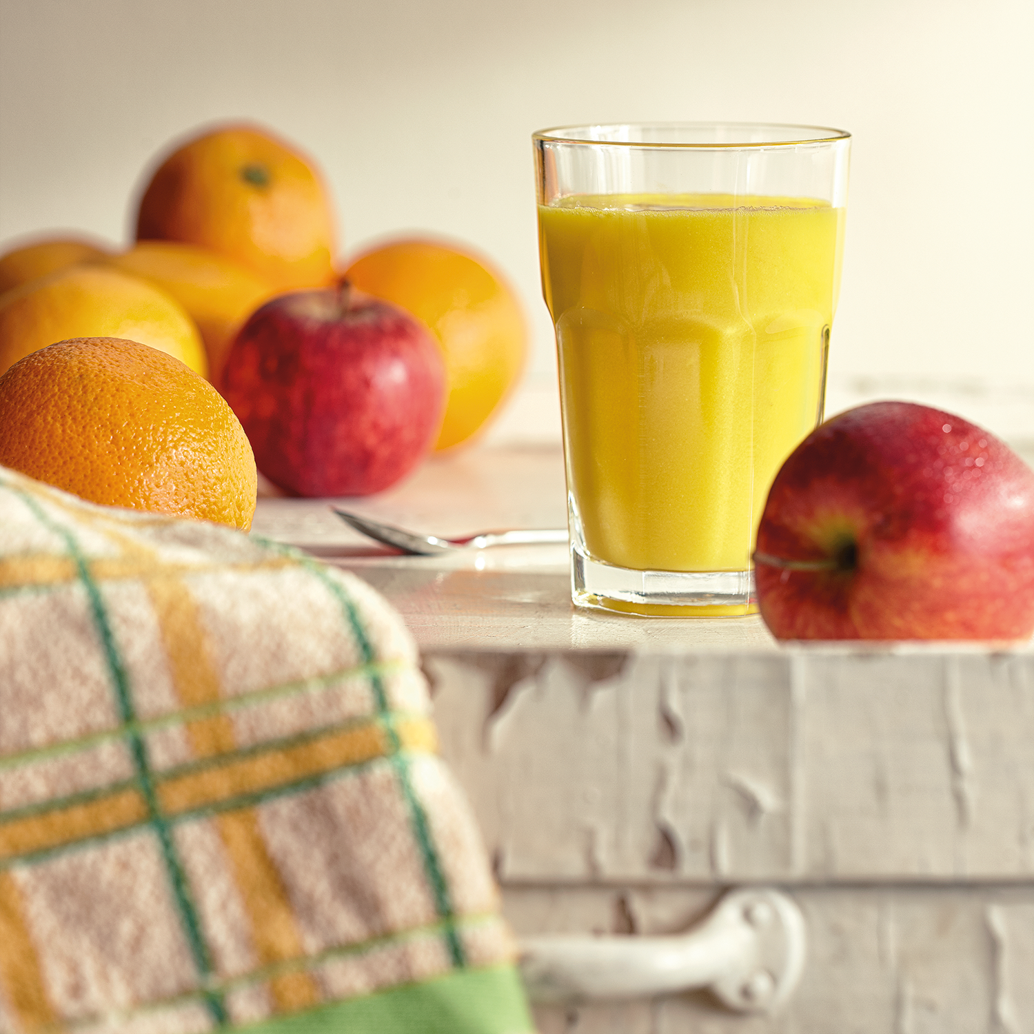 Stilleven: sinaasappelsap in glas, sinaasappels, rode appel en een handdoek op een wit oppervlak.