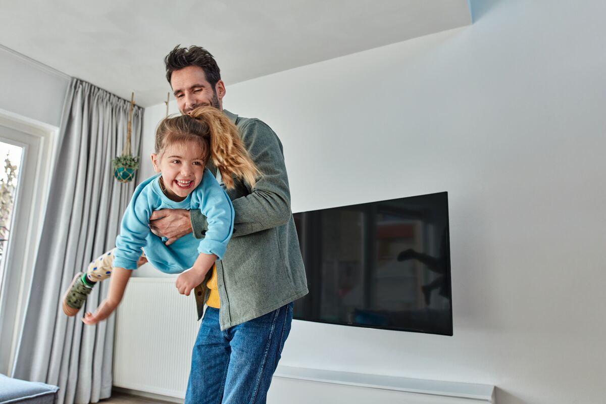 Un padre tiene in braccio la figlia che ride. C'è una TV sul muro.