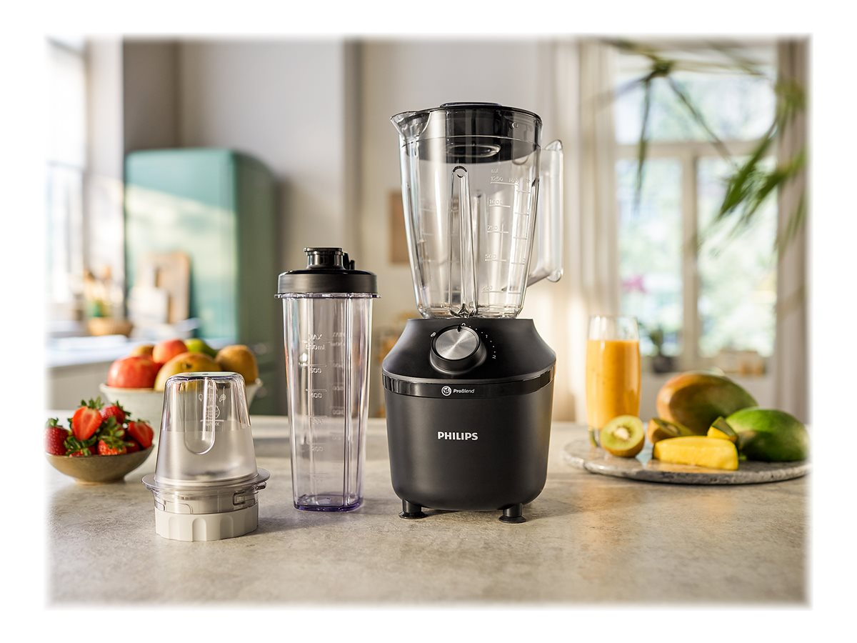 Mixer mit schwarzem Sockel, transparentem Behälter und Zubehör auf der Arbeitsplatte, Früchte im Hintergrund.