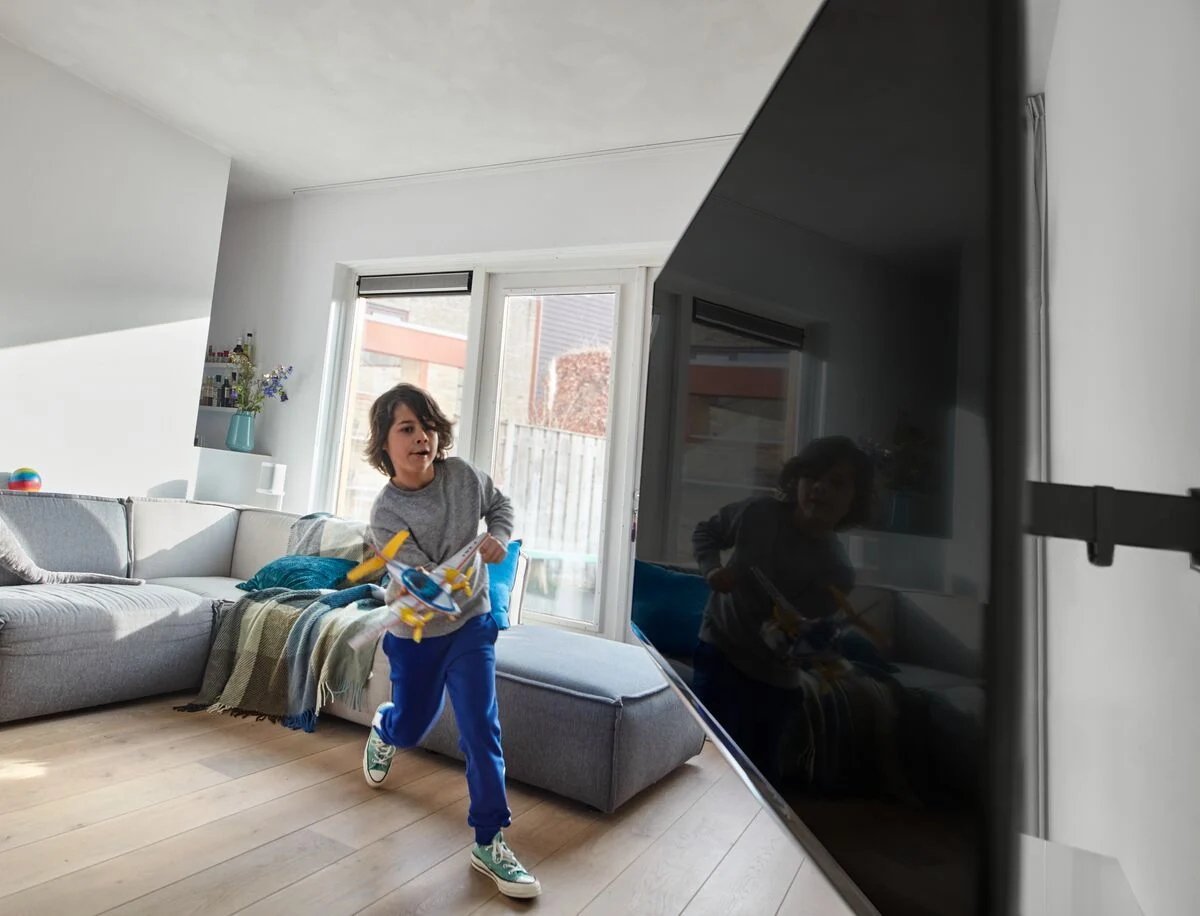 Junge rennt mit einem Spielzeugflugzeug im Wohnzimmer, Fernseher im Hintergrund.