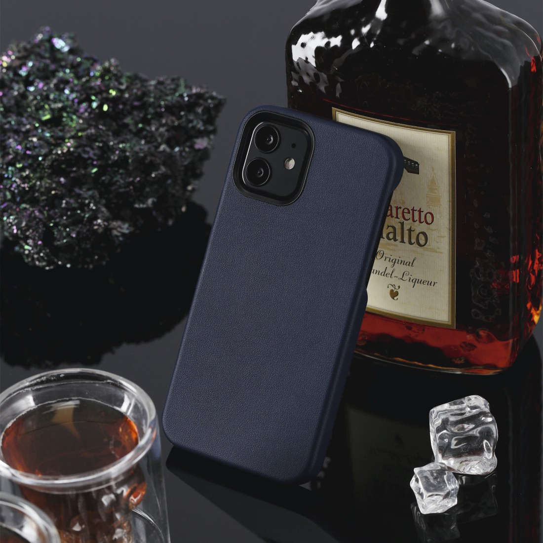 Dunkelblaue Handyhülle neben einer Likörflasche, einem Glas bernsteinfarbener Flüssigkeit und Eiswürfeln.