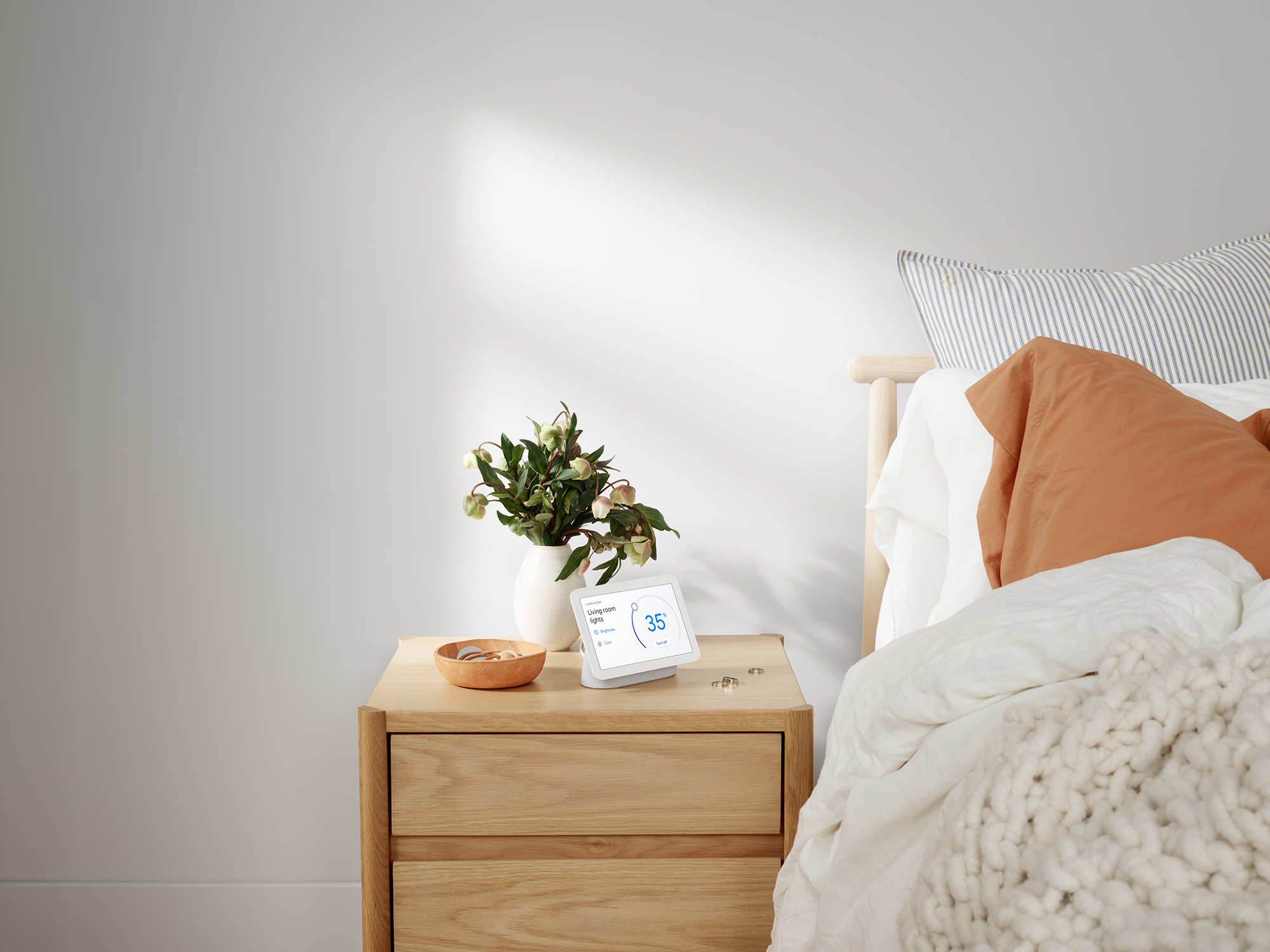 Nachtkastje met een slim display, bloemen en een kom erop. Bed en raam op de achtergrond.