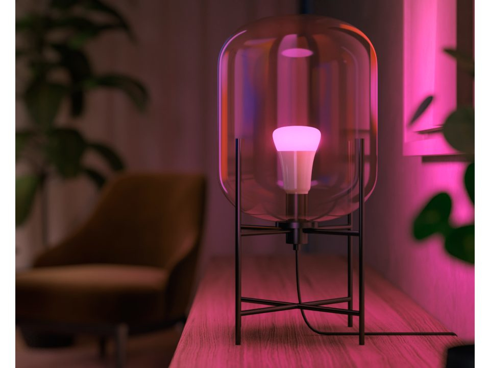A glass lamp with a pink lightbulb, on a wooden surface. A chair and a plant are in the background.