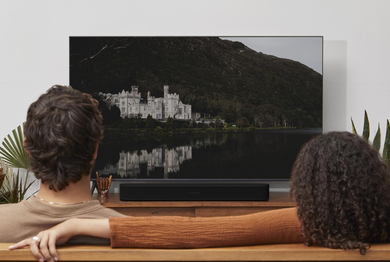 Zwei Personen auf einem Sofa schauen fern, eine Sonos Soundbar unter dem Fernseher, Landschaft.